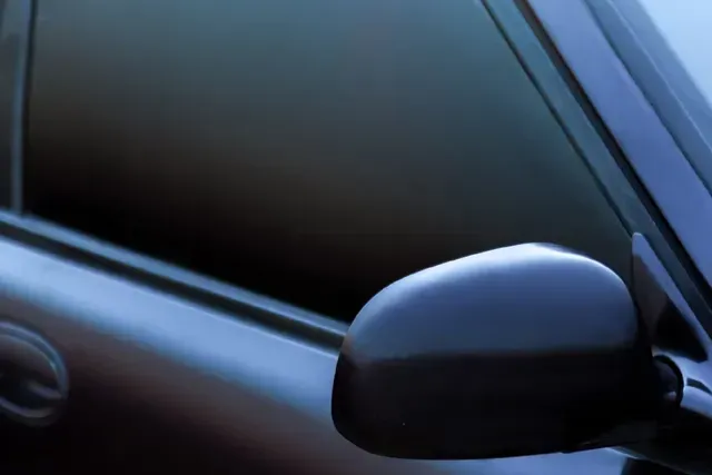 Primer plano del espejo retrovisor lateral de un coche negro contra una ventana tintada de azul.