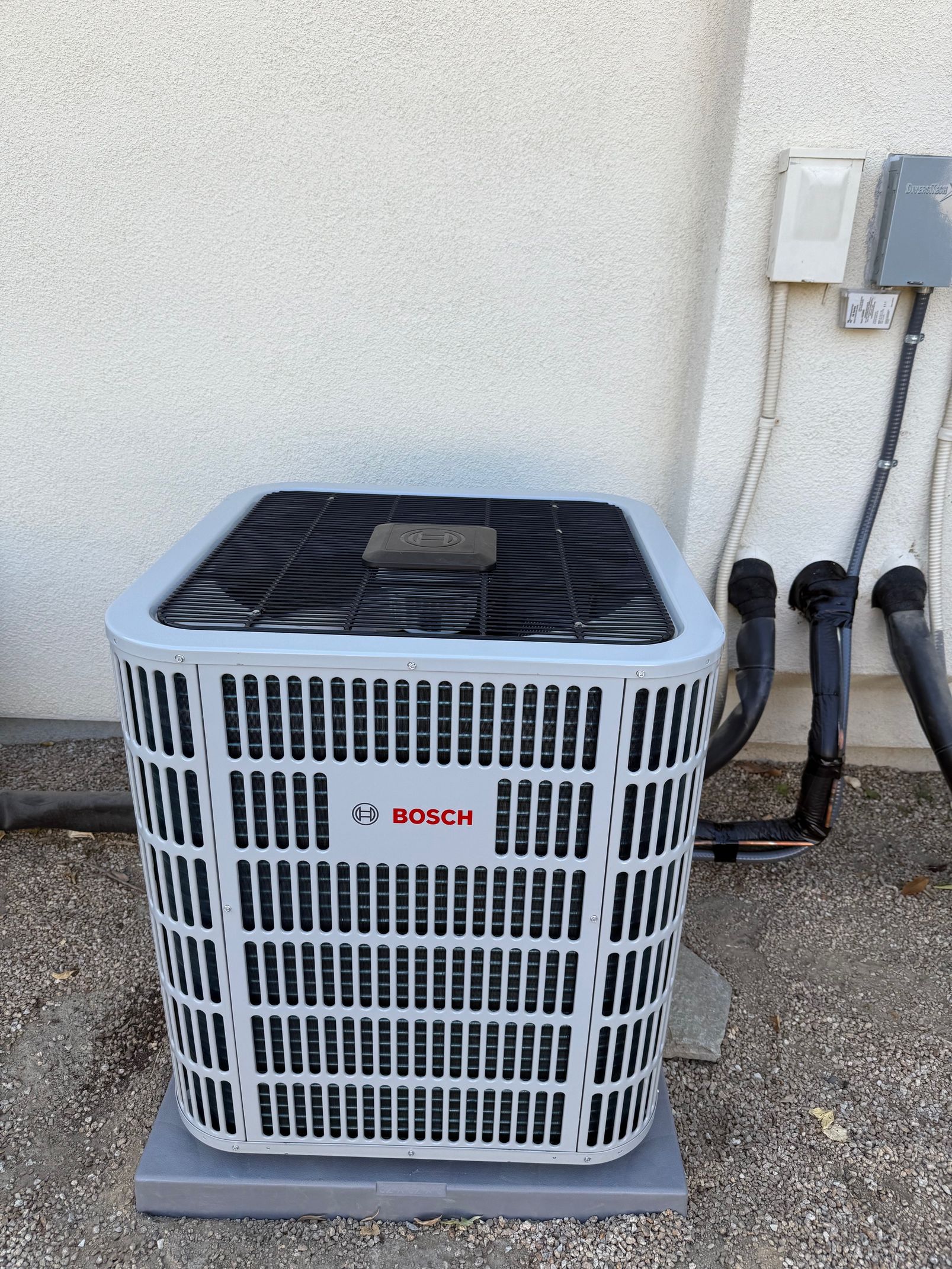 Bosch air conditioning unit, light gray and black, on a concrete pad near a stucco wall.