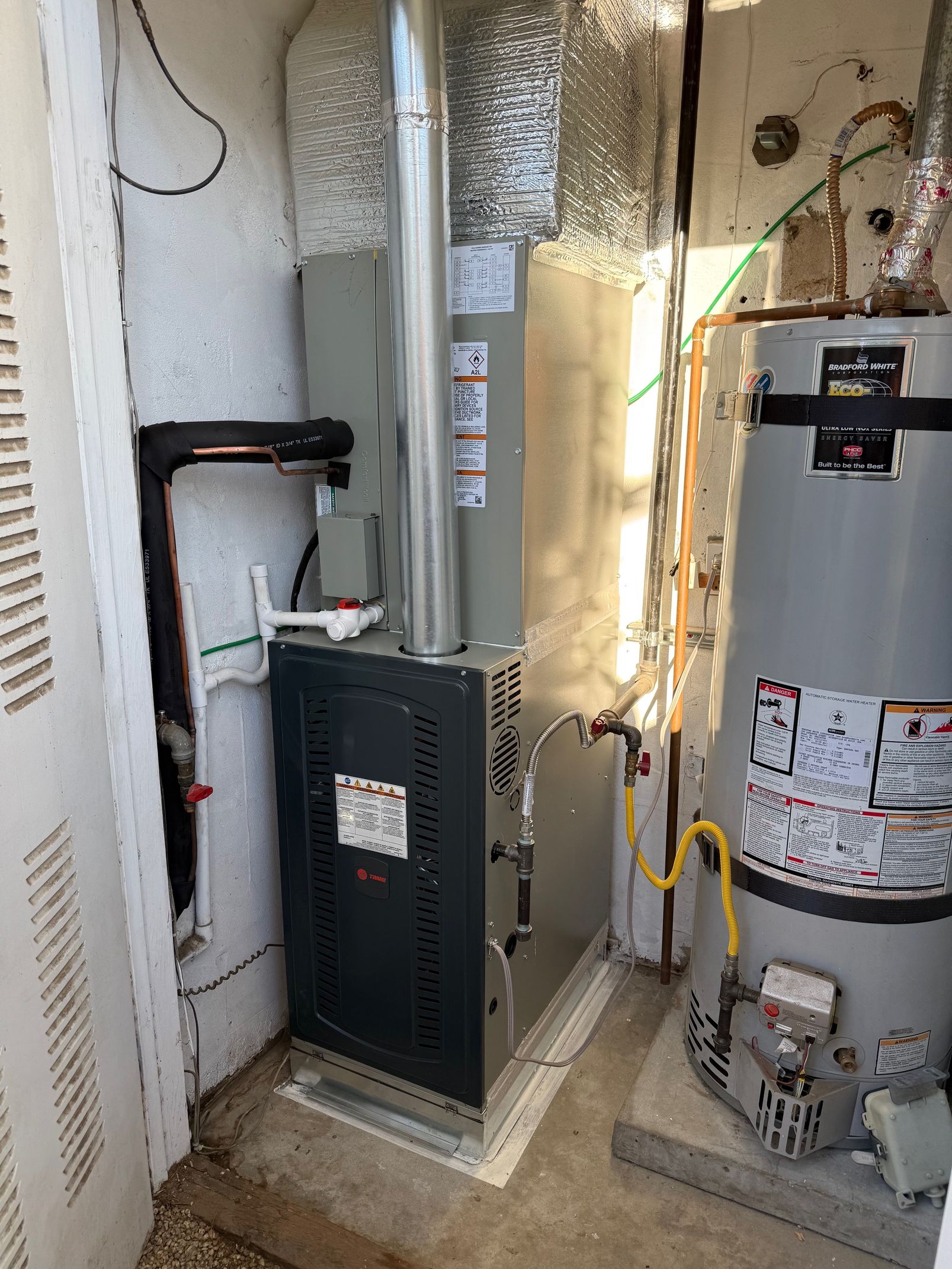 Furnace and water heater in a utility room. Metal ductwork, gray and black appliances, pipes.