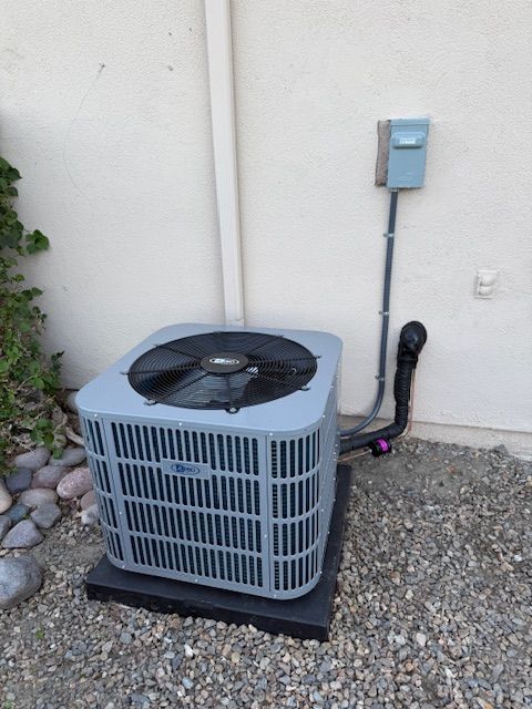 Outdoor air conditioning unit with black fan, against a beige wall, gravel ground.