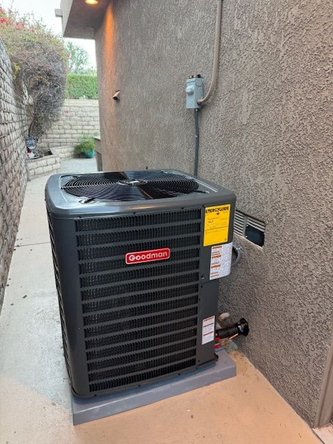 Goodman air conditioner unit on a concrete pad next to a stucco wall, electrical box visible.