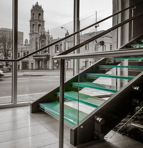 Modern glass staircase with blue-tinted steps, steel frame, and city view through window.