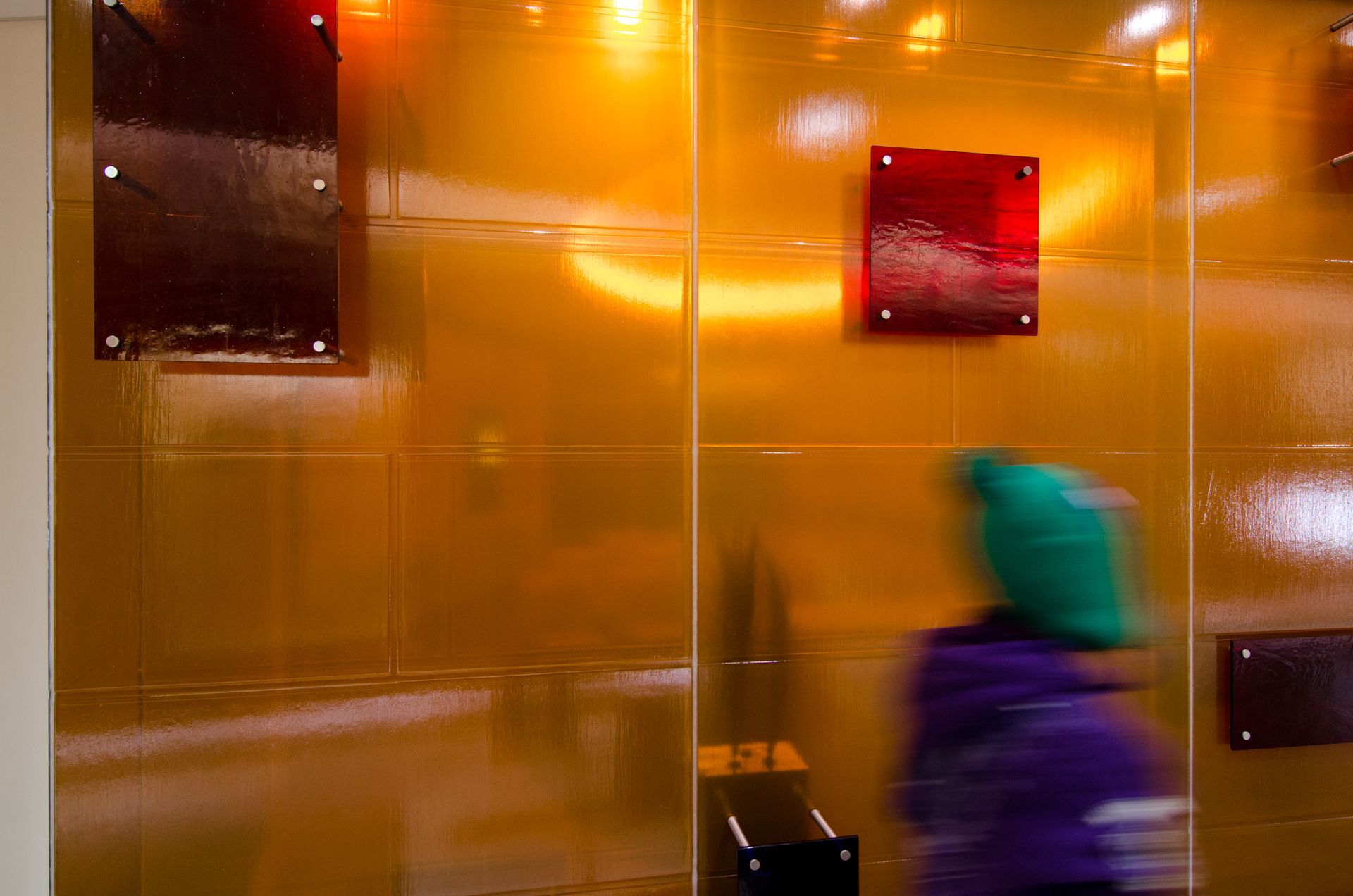 Blurred person in teal hood and purple jacket walking past a shiny orange wall with dark panels.