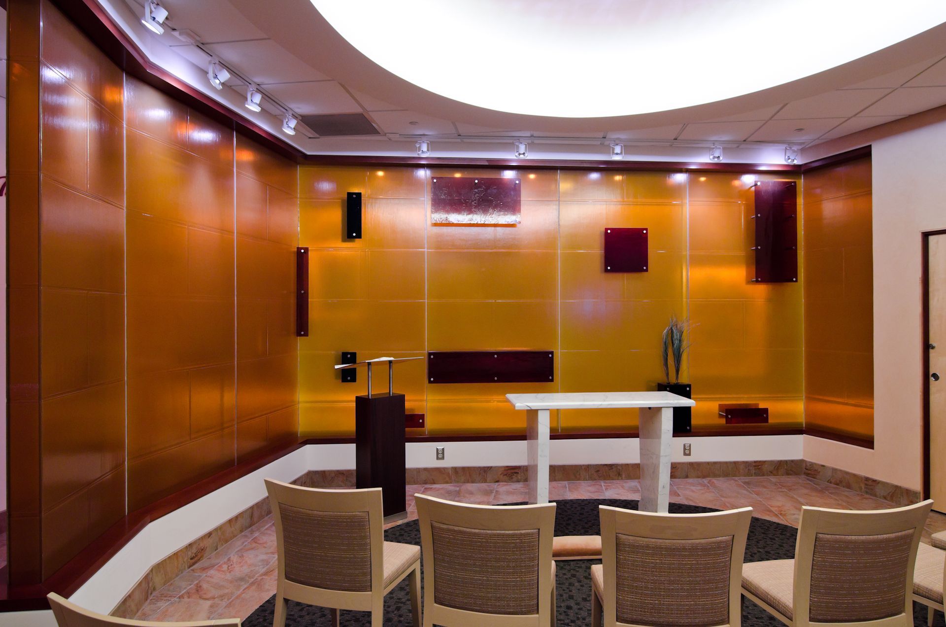 Modern lounge with warm wood panels, recessed lighting, and rows of chairs facing a small table at the front.