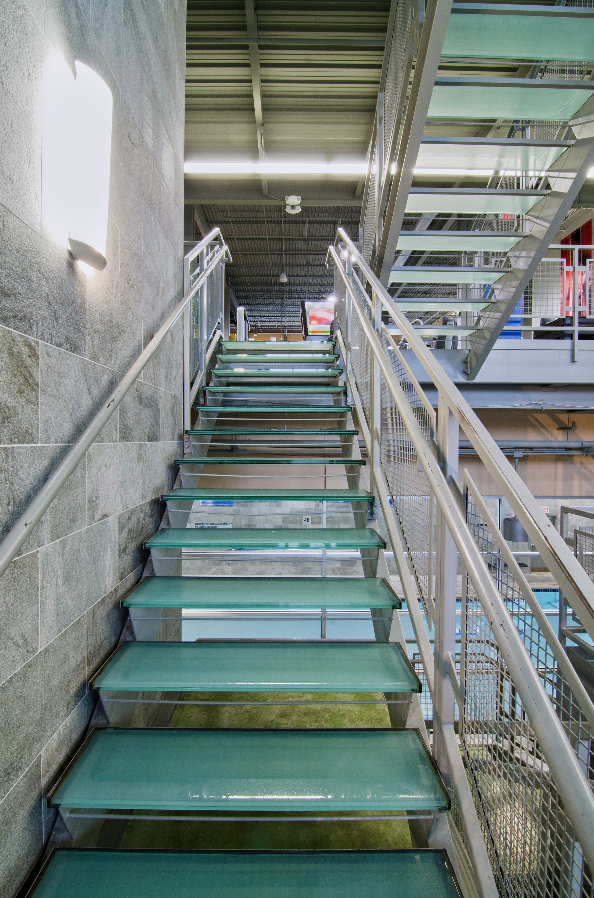 Narrow green metal staircase under an overhang with railings and chain-link fencing, viewed from below