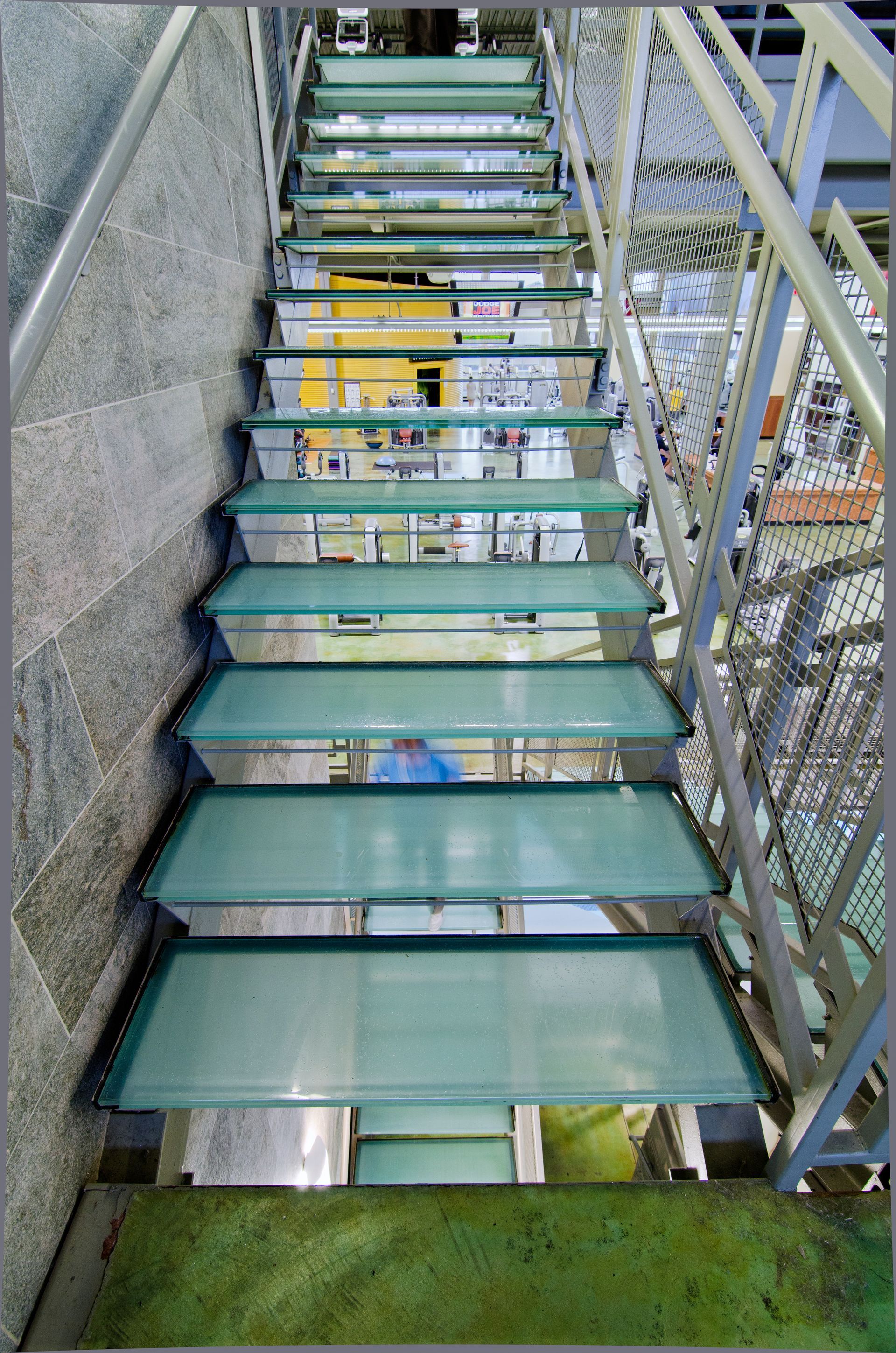 Outdoor staircase with glass steps, metal railings, and green turf below beside a chain-link fence