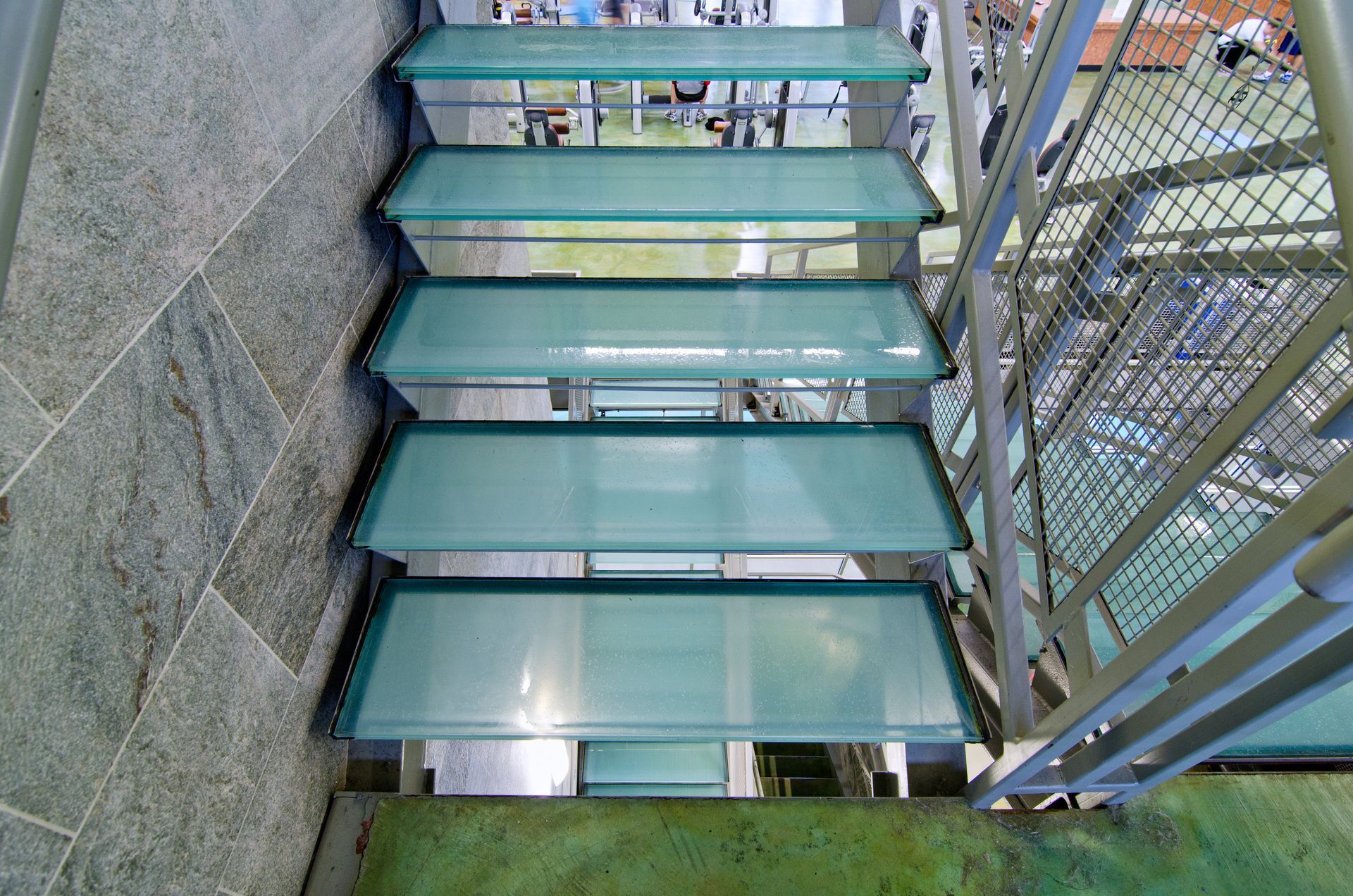 Glass steps on a narrow outdoor staircase beside a metal fence and stone wall.