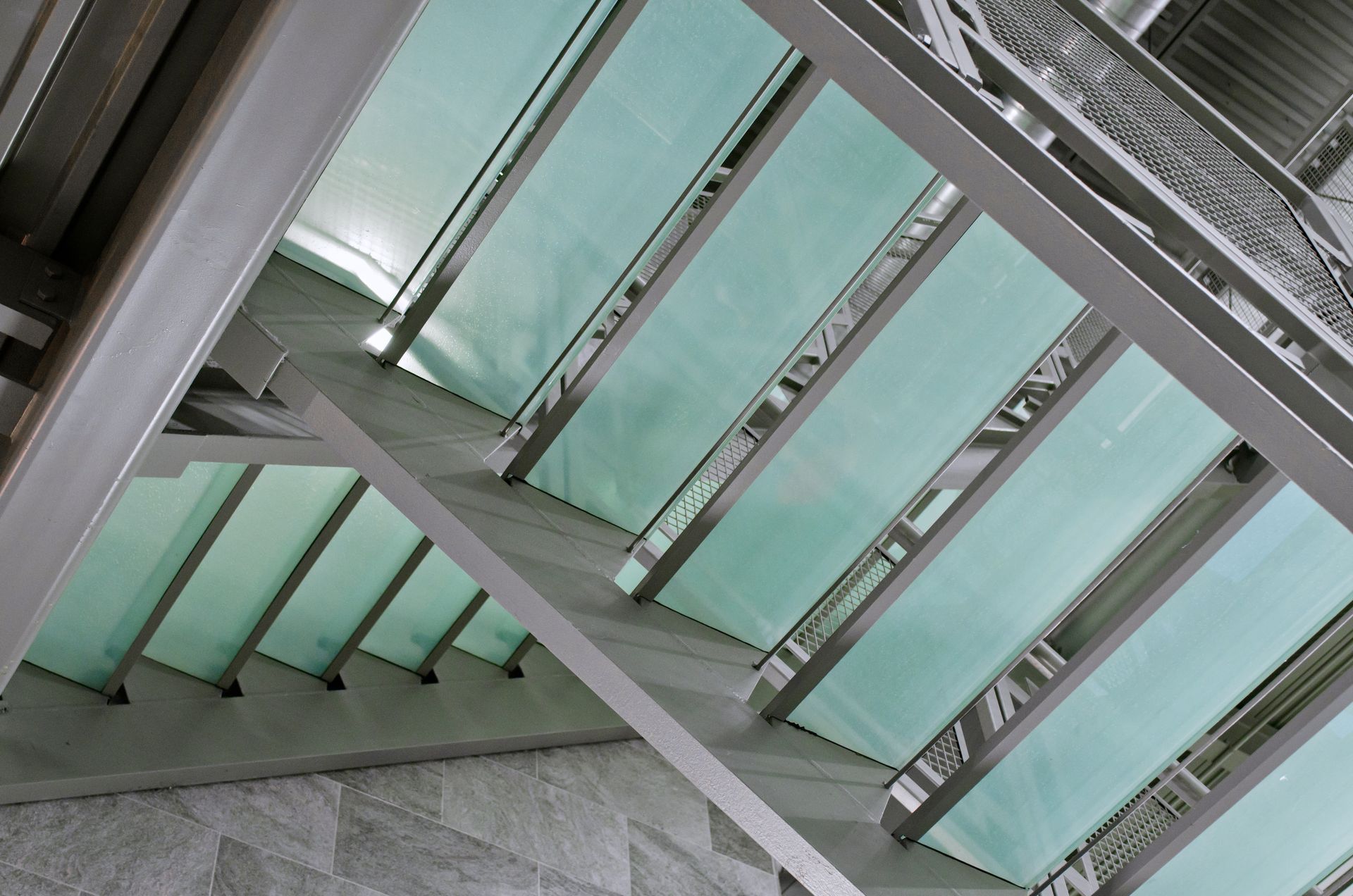 Glass stairwell with mint-green panels and metal beams seen from below