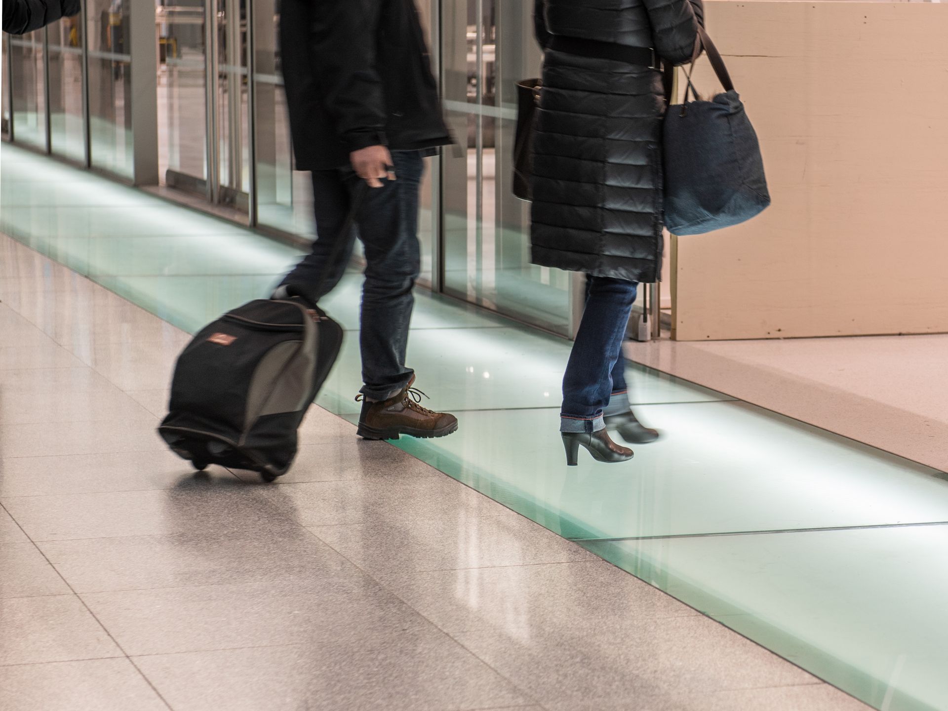 Two travelers walking in an airport corridor, one pulling a black rolling suitcase and one carrying a tote bag