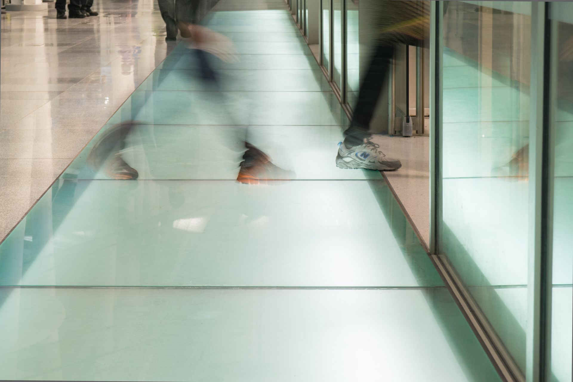 Blurry legs walking through a bright glass-walled corridor with a reflective floor
