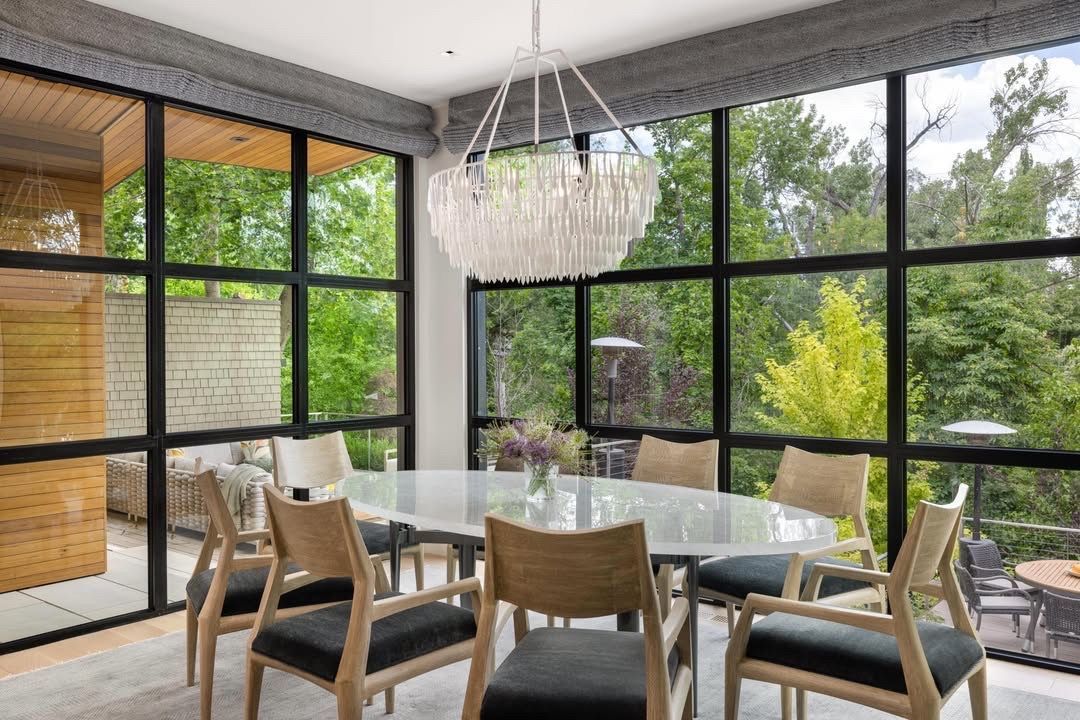 Modern dining room with glass table, light wood chairs, and floor-to-ceiling windows overlooking greenery