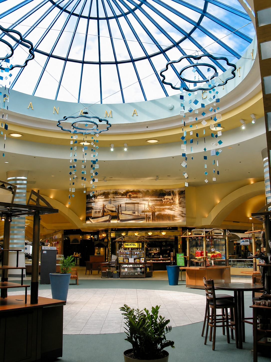 Indoor shopping mall with a glass dome ceiling, hanging blue decorations, and storefronts below