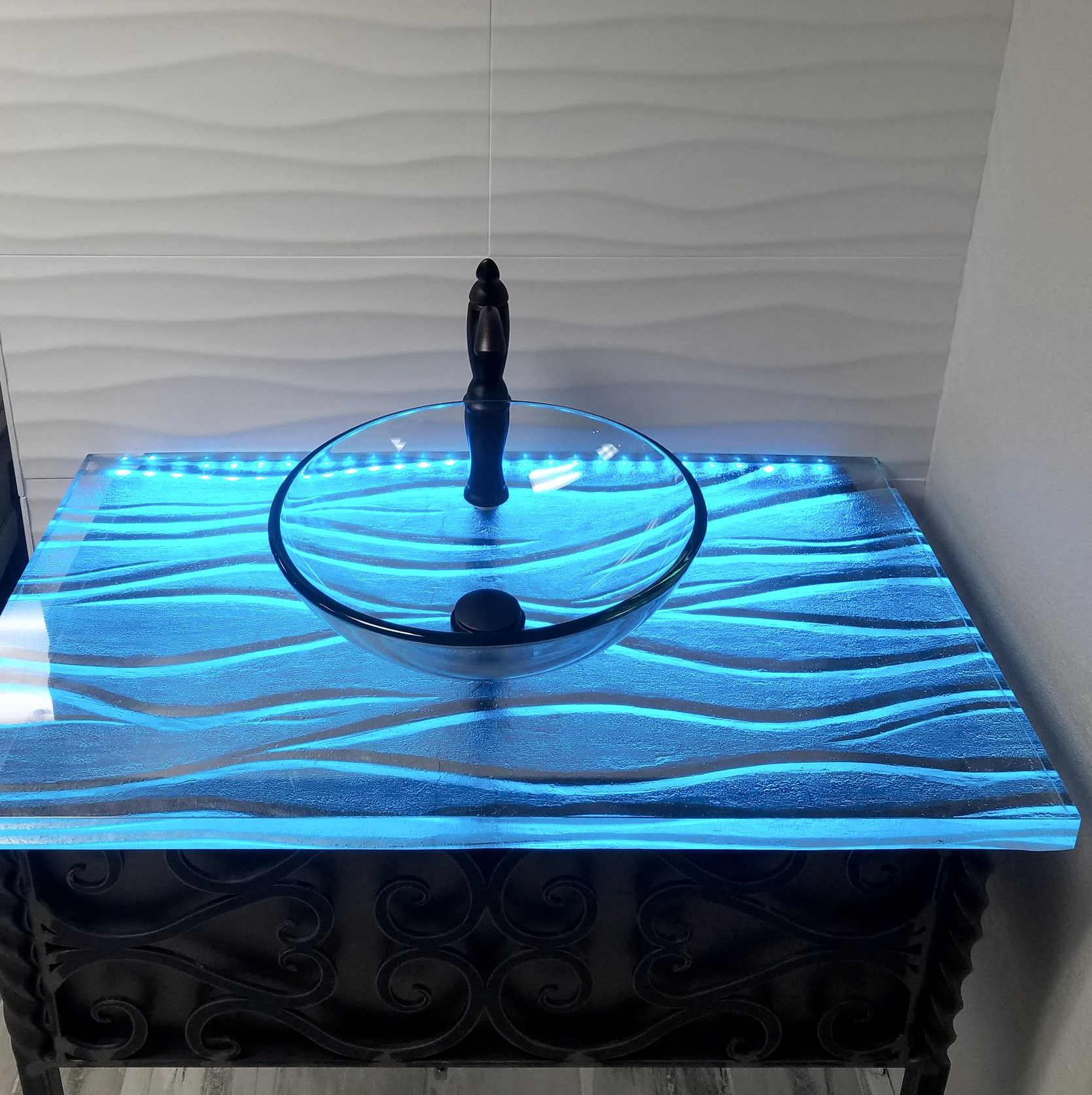 Vanity with illuminated blue waves, glass sink, black faucet, set against a textured white wall.