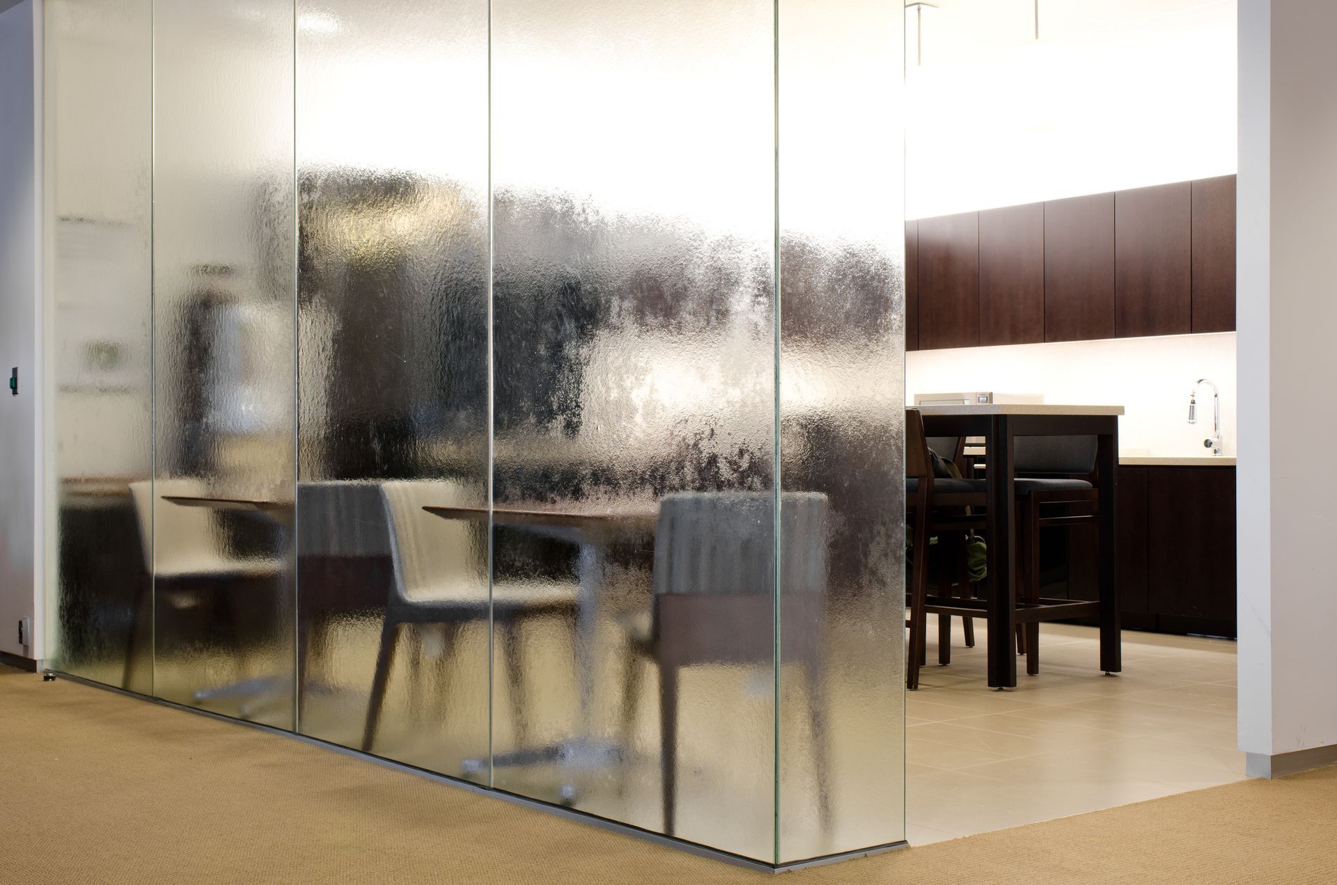 Modern office lounge with frosted glass walls, chairs, and a kitchenette area