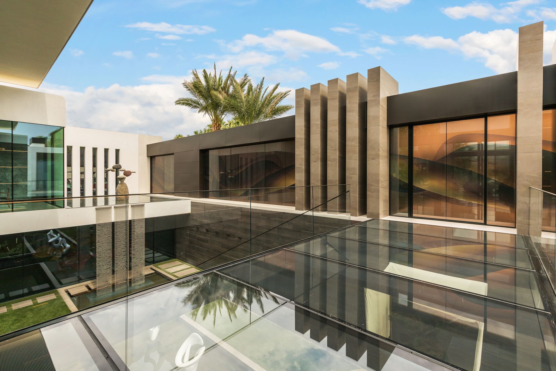 Modern luxury house with glass walls, stone columns, and a reflective pool under a partly cloudy sky