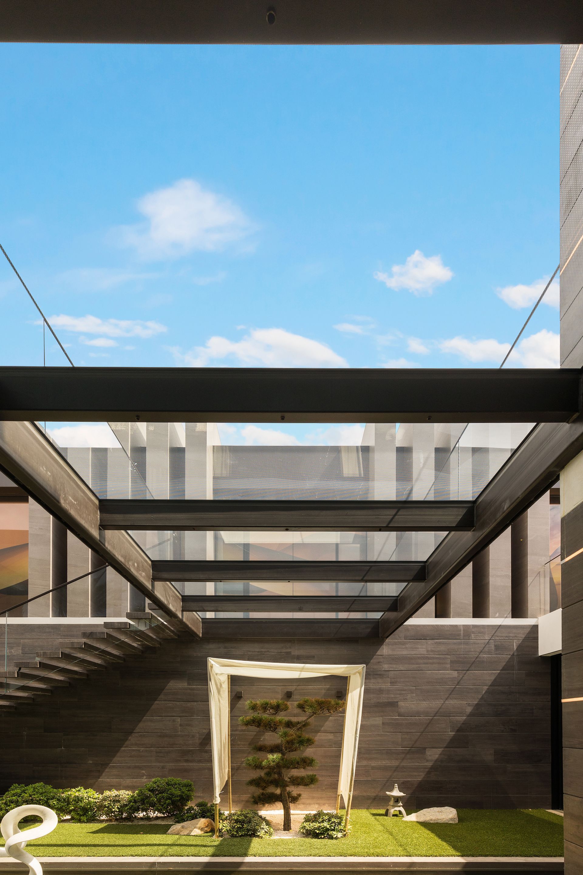Modern courtyard with stone walls, glass canopy, and a small tree under blue sky.