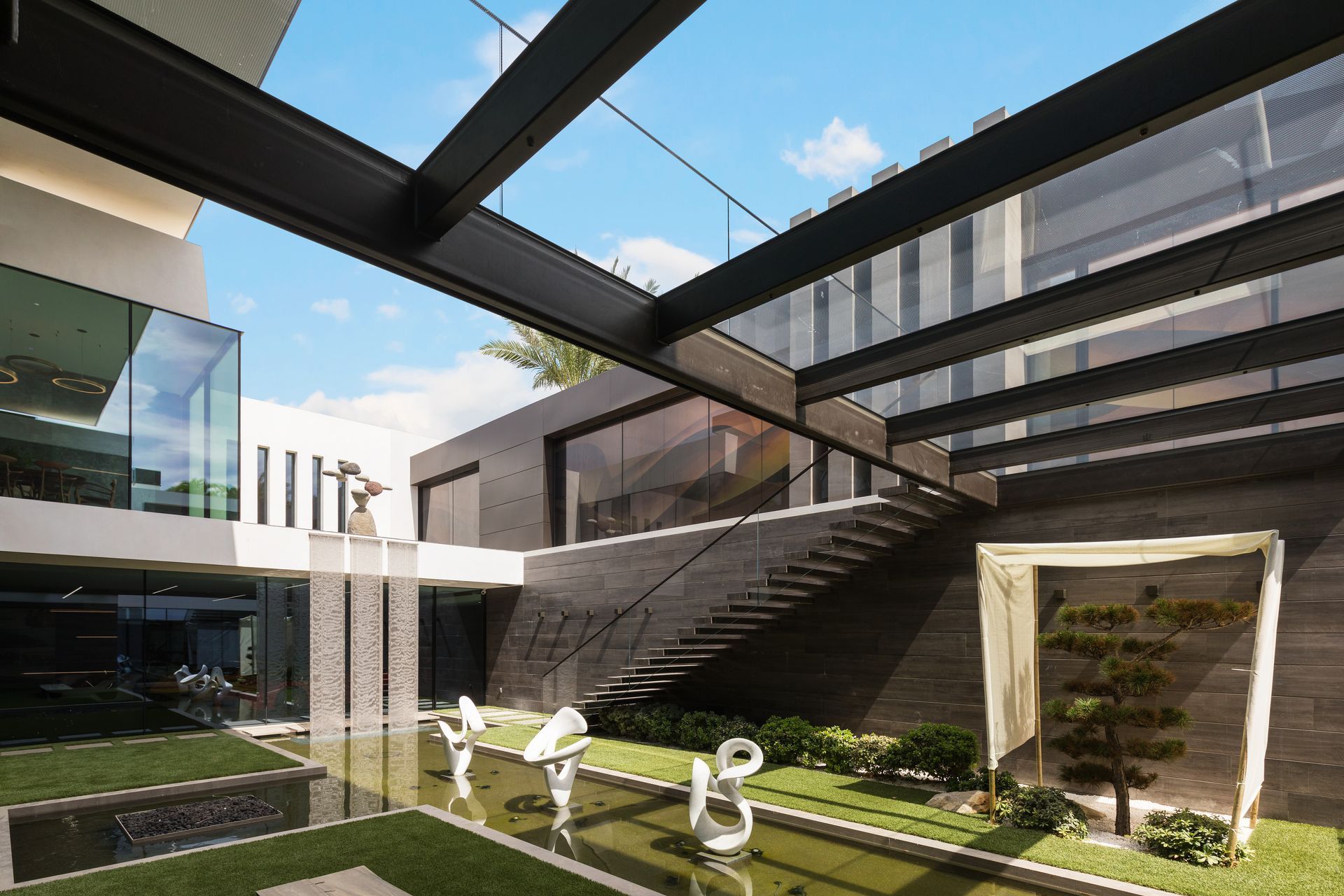 Modern courtyard with white sculptures, glass buildings, steps, and a blue sky overhead