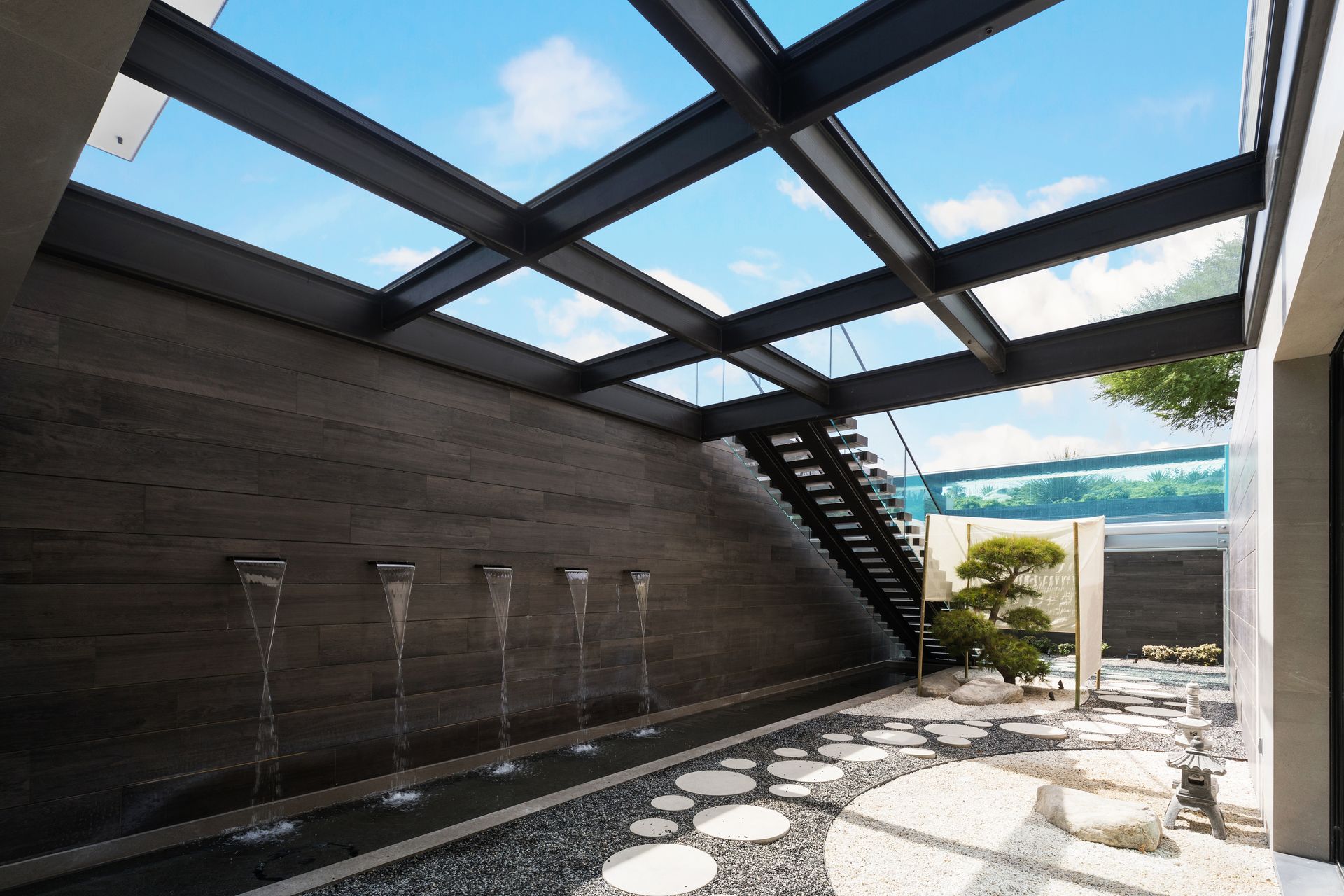 Modern courtyard with wall fountains, stone path, and glass roof framing blue sky