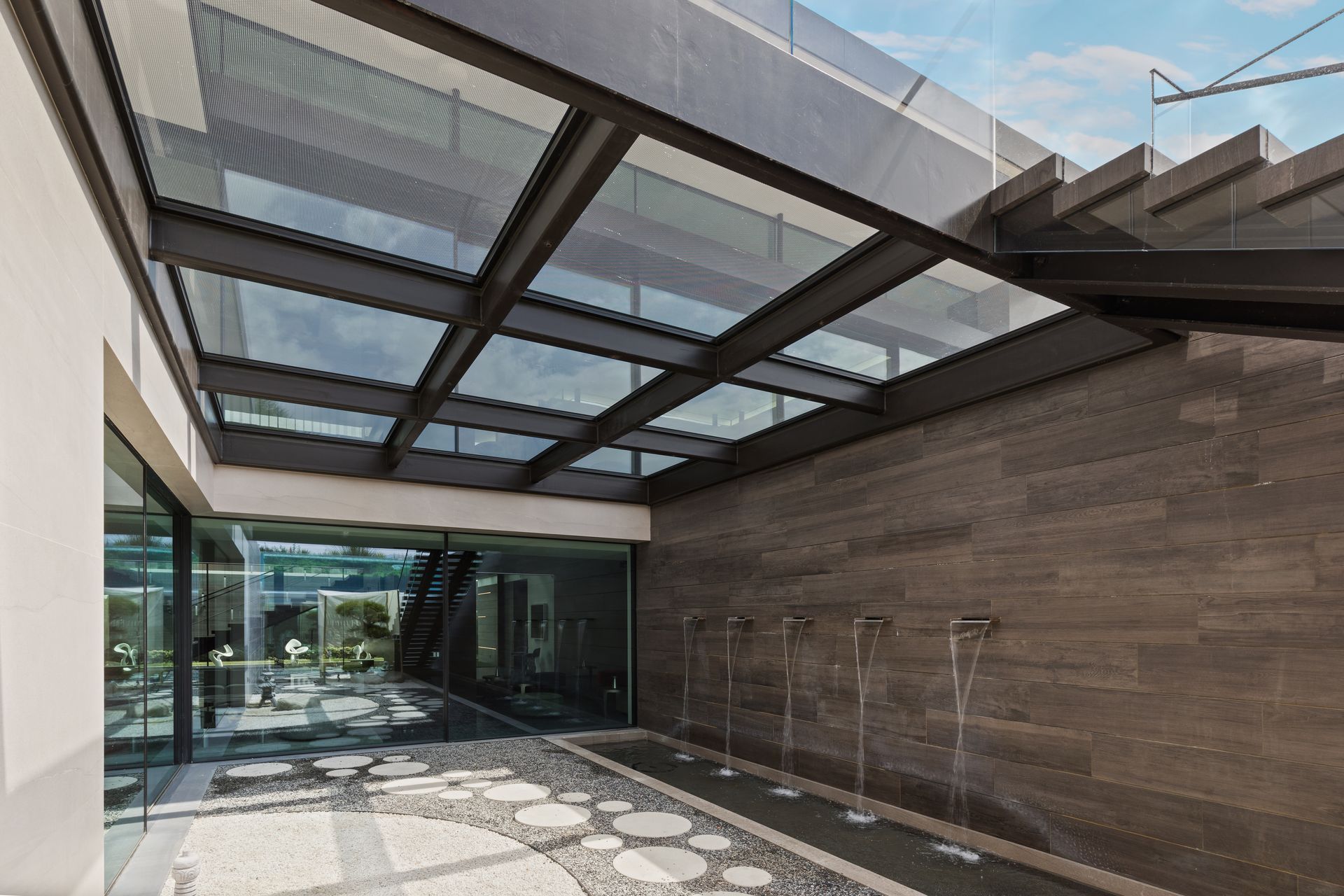 Modern covered courtyard with glass roof, stone walls, and patterned floor leading to a glass entrance.