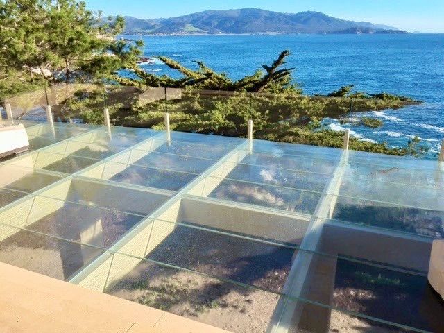 Glass-floored outdoor platform overlooking ocean with trees and mountains in the background.
