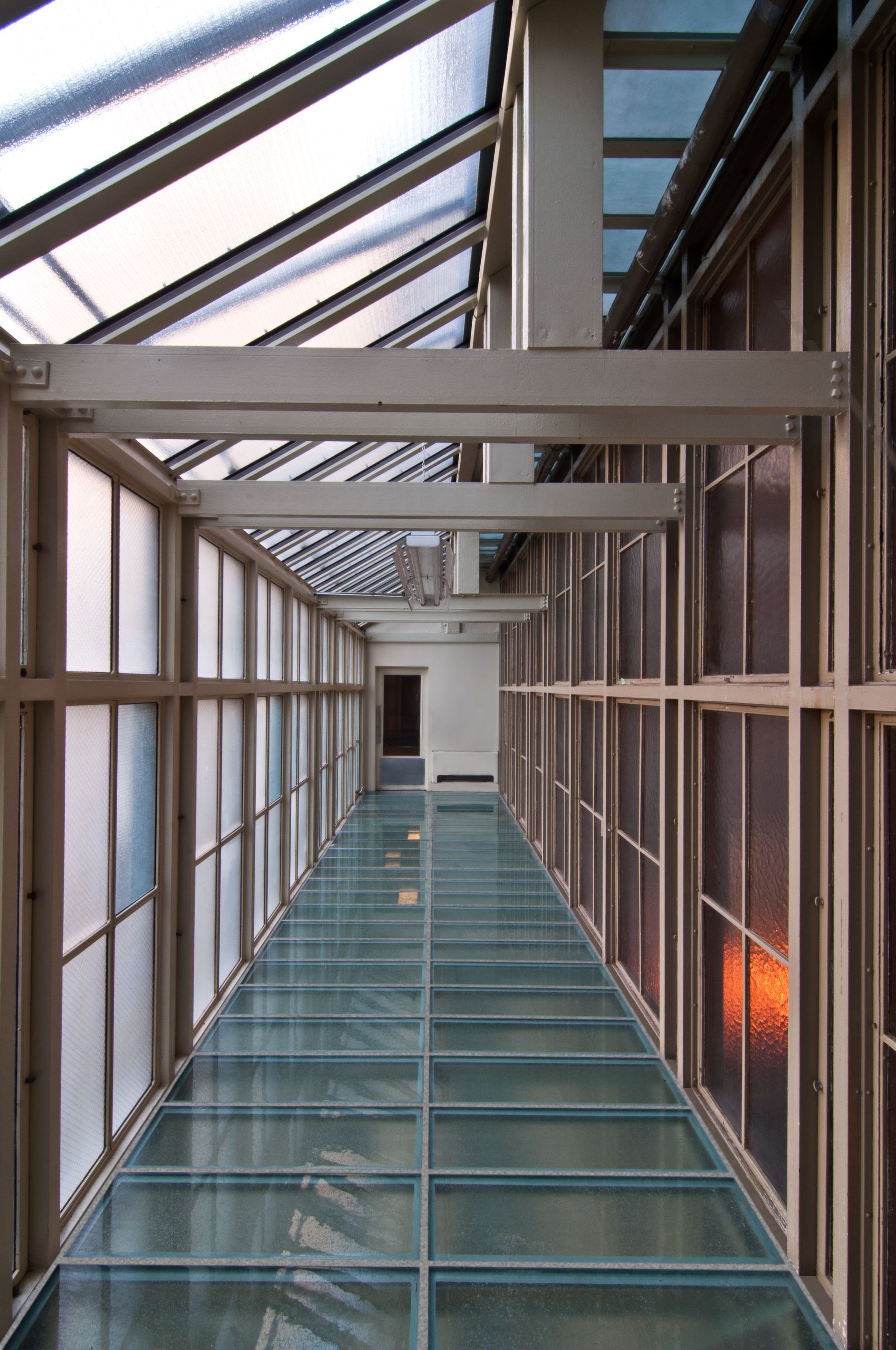 Glass-enclosed corridor with metal beams and a teal floor, lit by daylight.