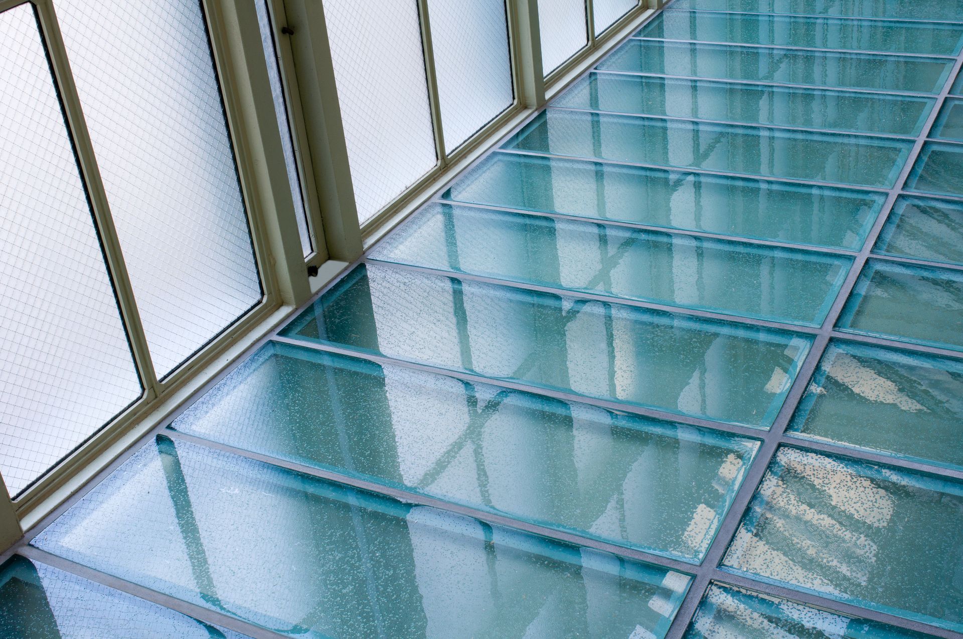 Blue glass floor with metal grid panels beside tall windows, viewed in perspective