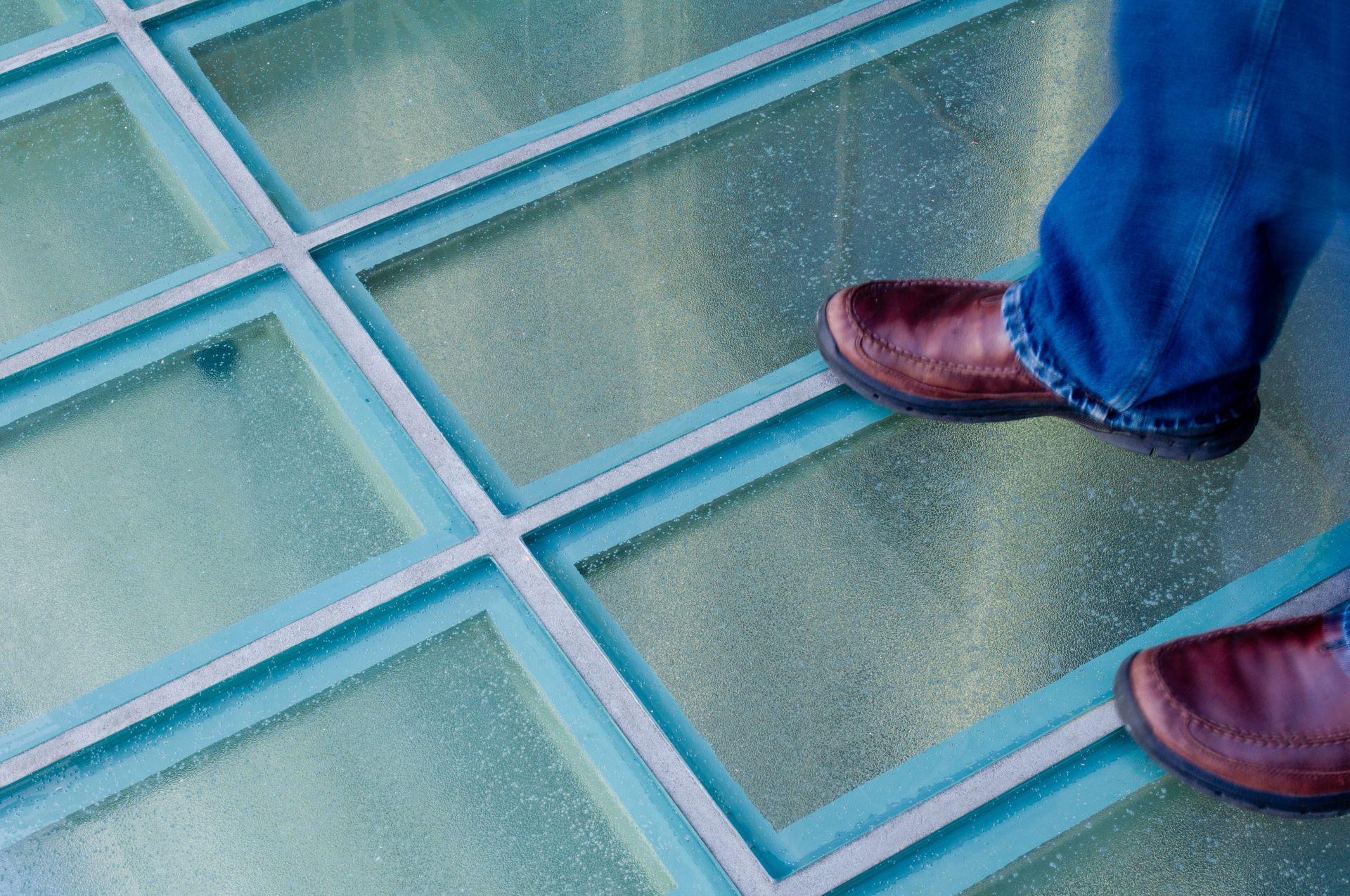 Two brown shoes on a blue glass-tiled floor with a person in jeans standing nearby
