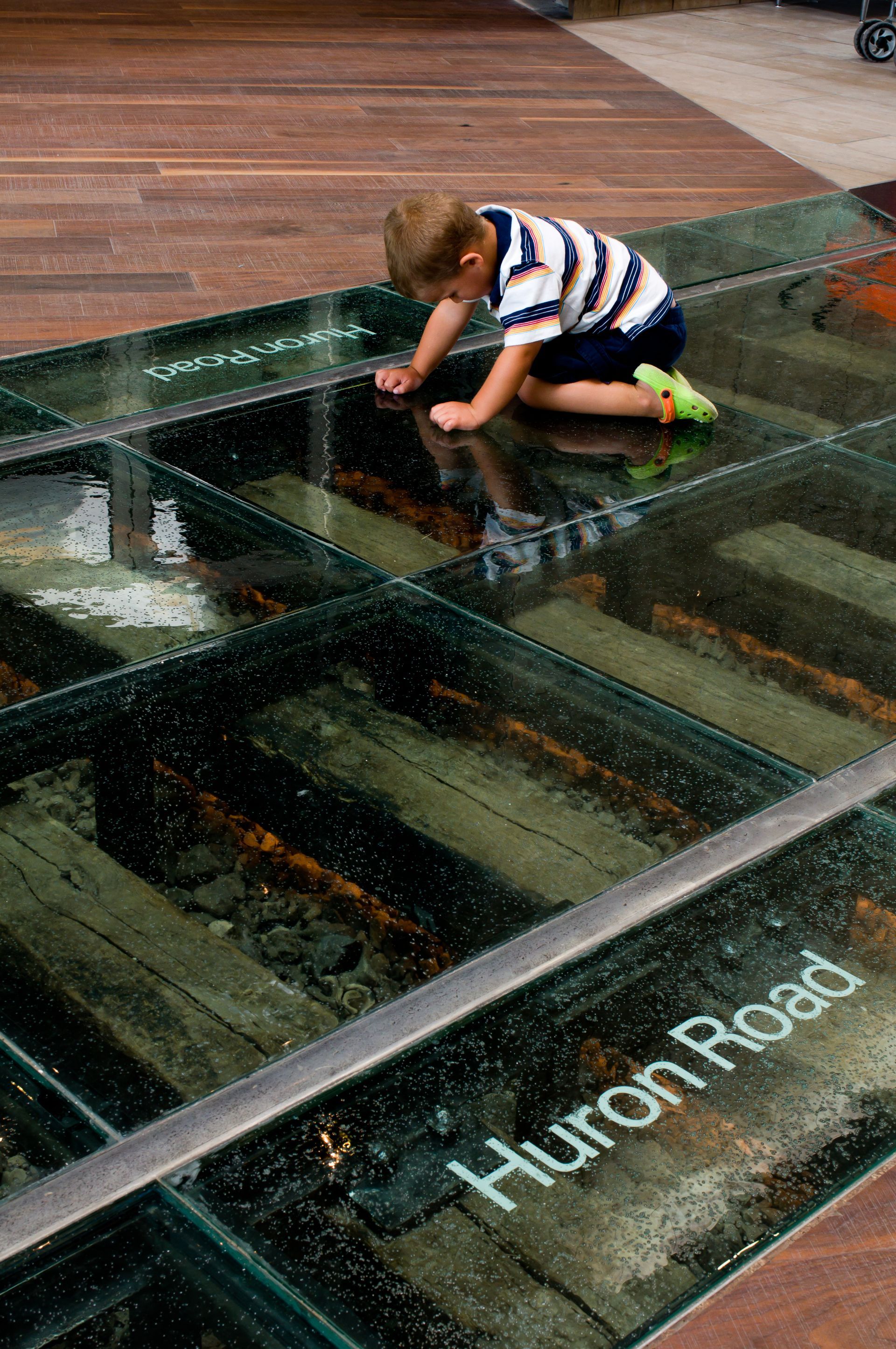 Boy crouching on glass floor panels with water below at Huron Road exhibit.