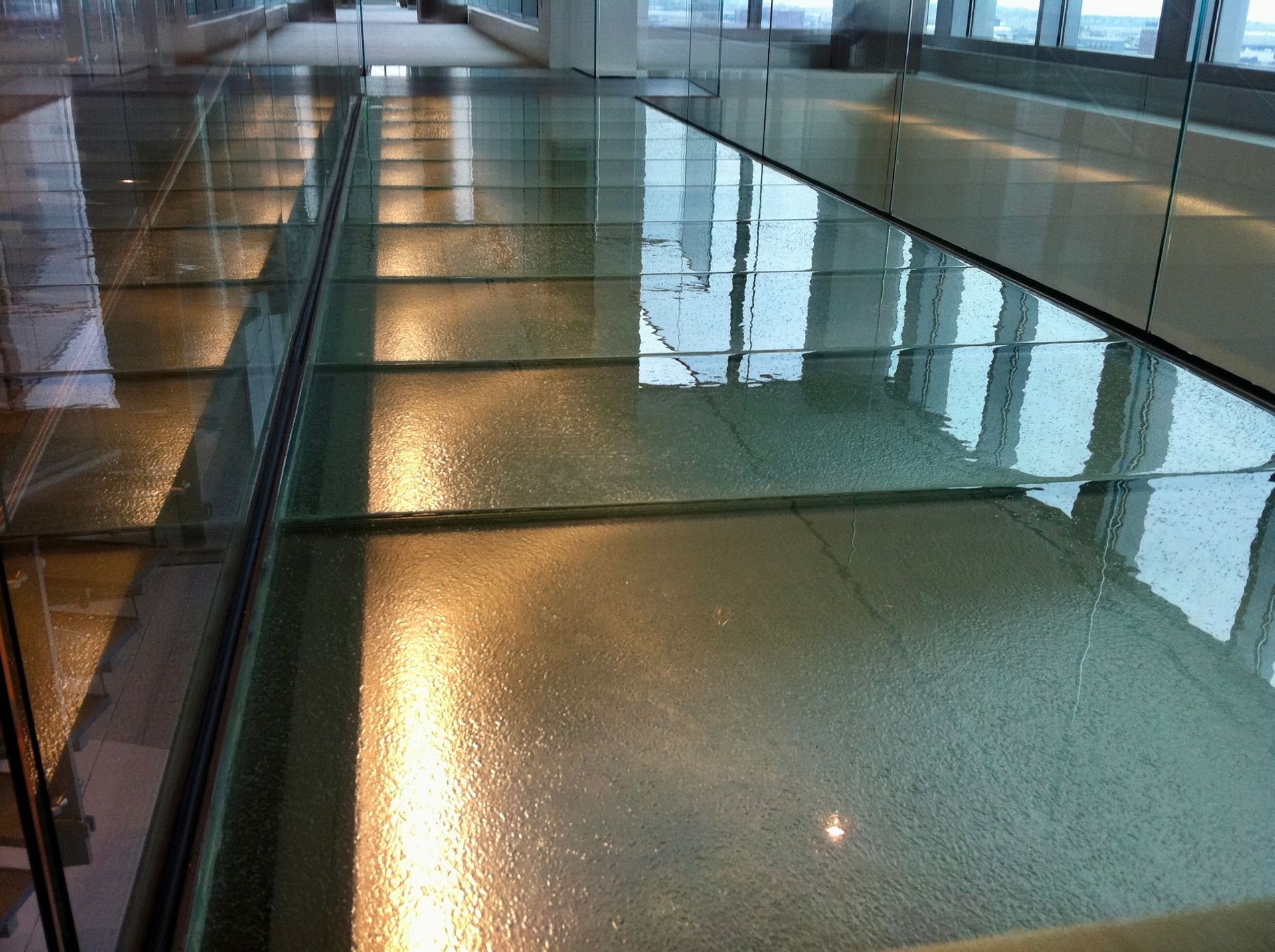 Glass walkway with reflections and a metal railing in a bright indoor hallway