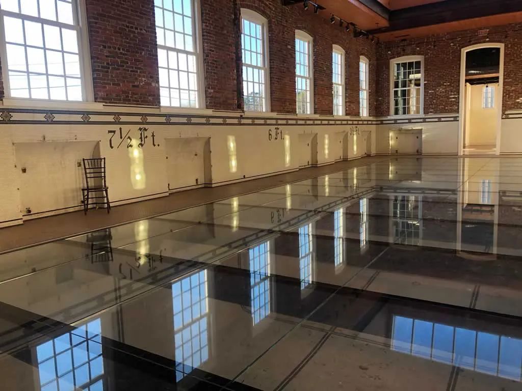 Indoor pool with brick walls and a reflective glass floor, featuring tall windows and a doorway.