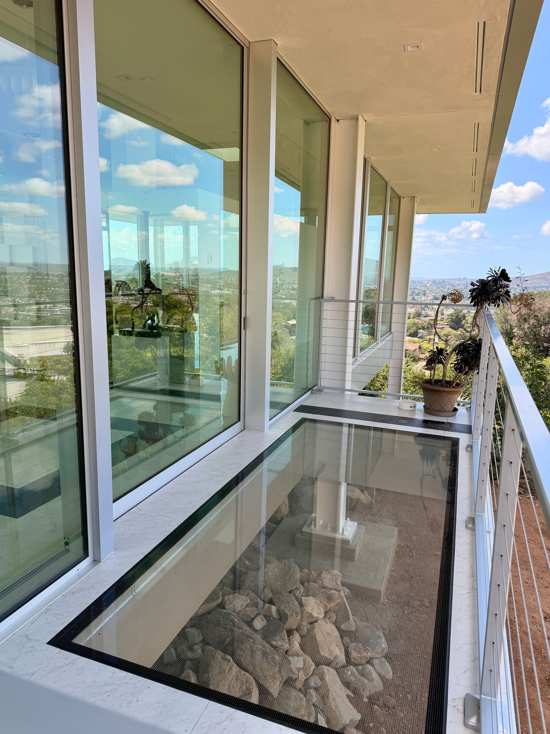 Balcony with glass railing, beige walls, and large windows overlooking a sunny outdoor view. Large glass floor