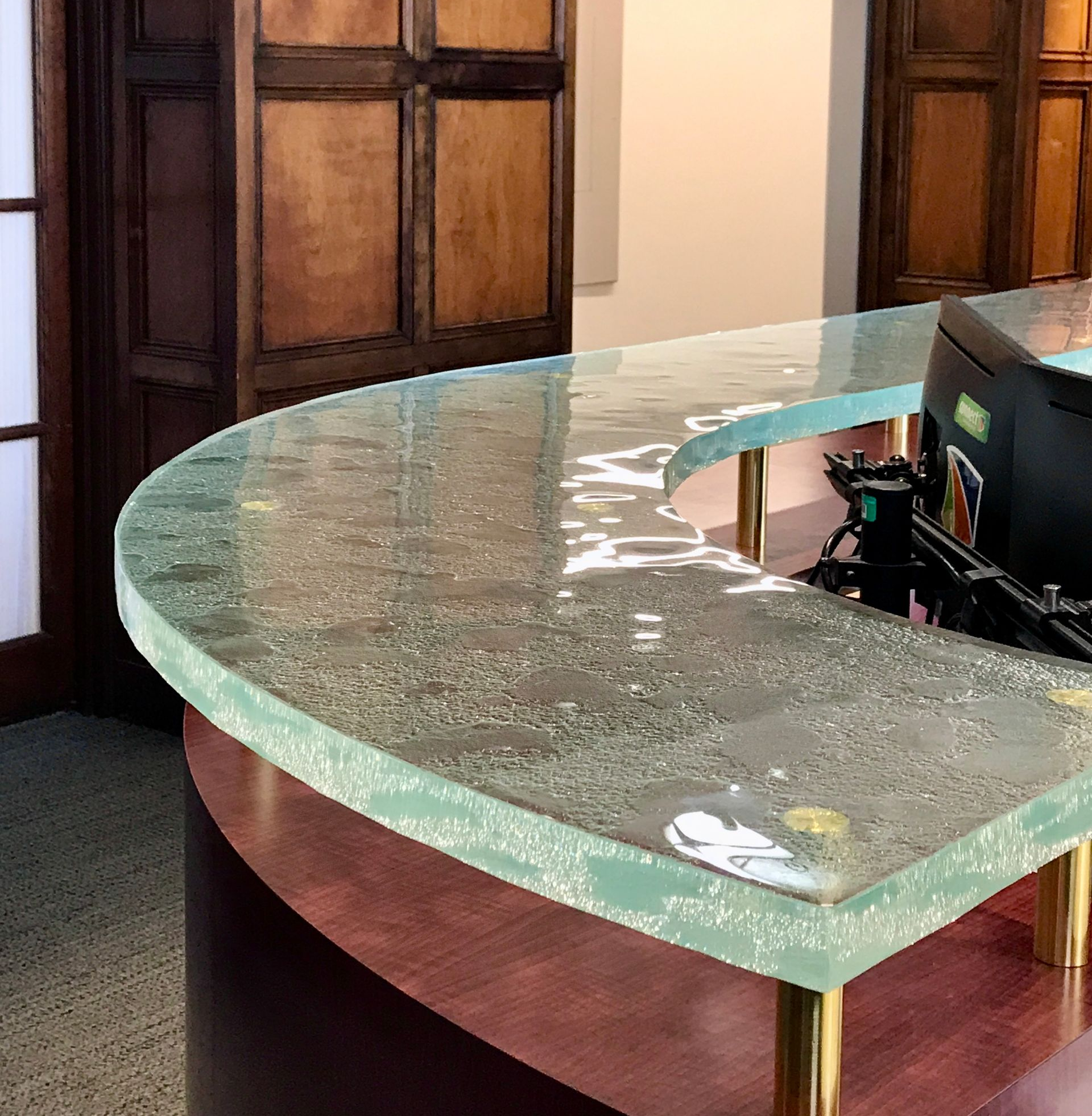 Curved, frosted glass countertop on a dark wood and gold leg reception desk. Wooden doors in the background.