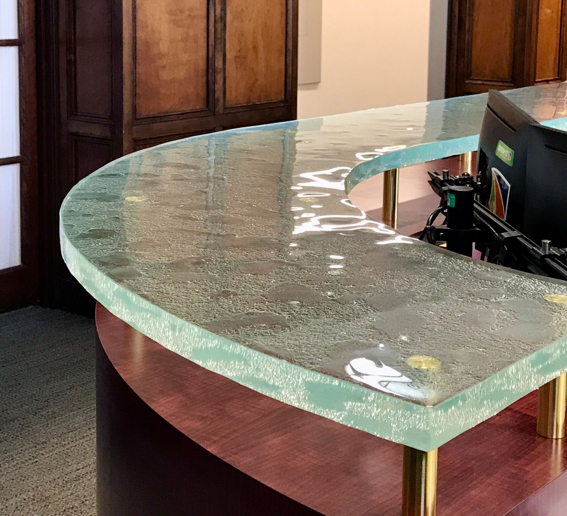 Curved reception desk with clear textured glass top, red wood base, and gold legs, in a room with wood paneling.