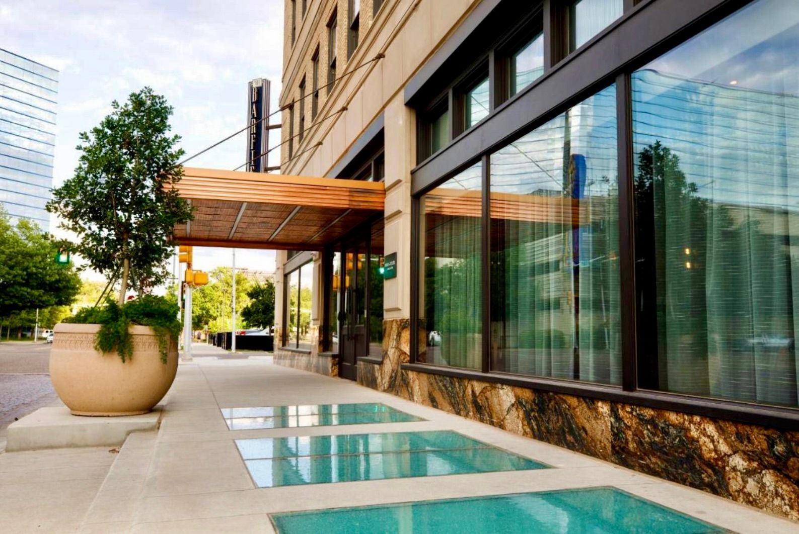 Exterior sidewalk with glass skylights, a large potted tree, and a modern building entrance under a wood-paneled awning.