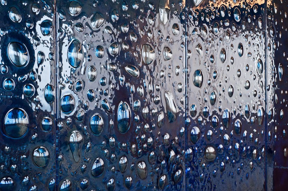 Rain droplets on a blue glass surface with blurred lights behind it