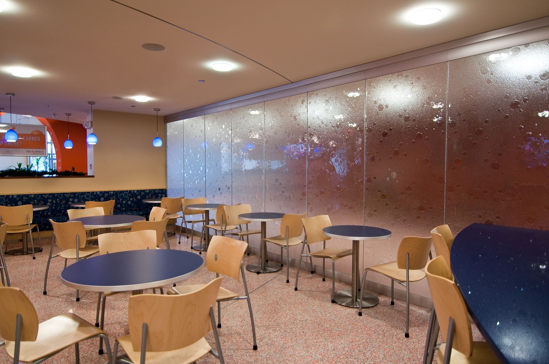Empty café seating with round tables and light wood chairs beside a large reflective wall.