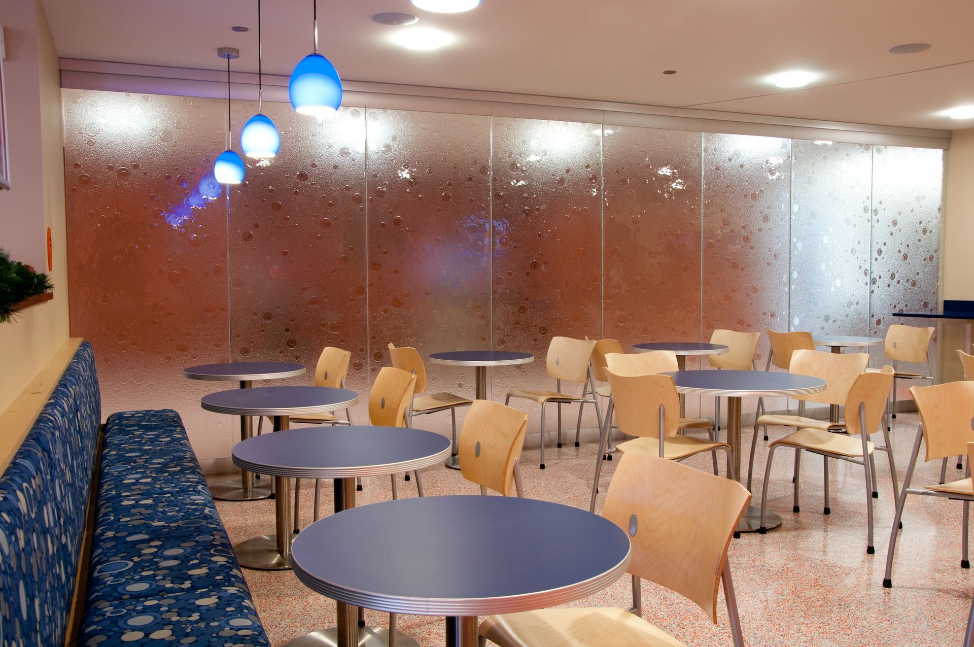 Empty cafe with round tables, wooden chairs, blue pendant lights, and a mosaic wall.