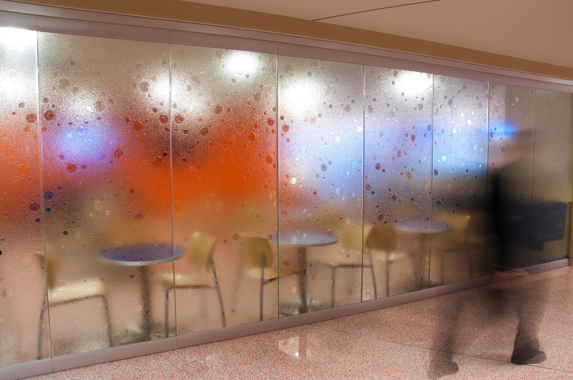 Blurred person walking past colorful frosted glass wall with café tables and chairs