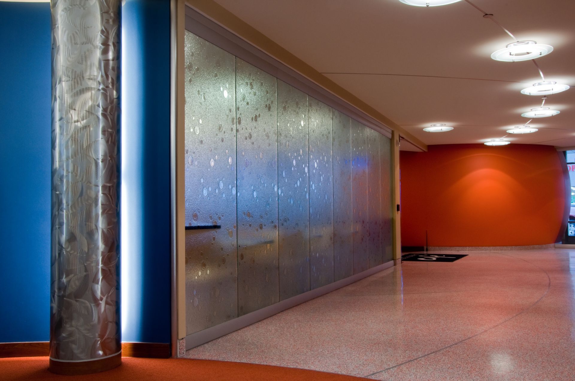 Empty modern hallway with orange wall, blue accents, frosted glass partition, and reflective metal column