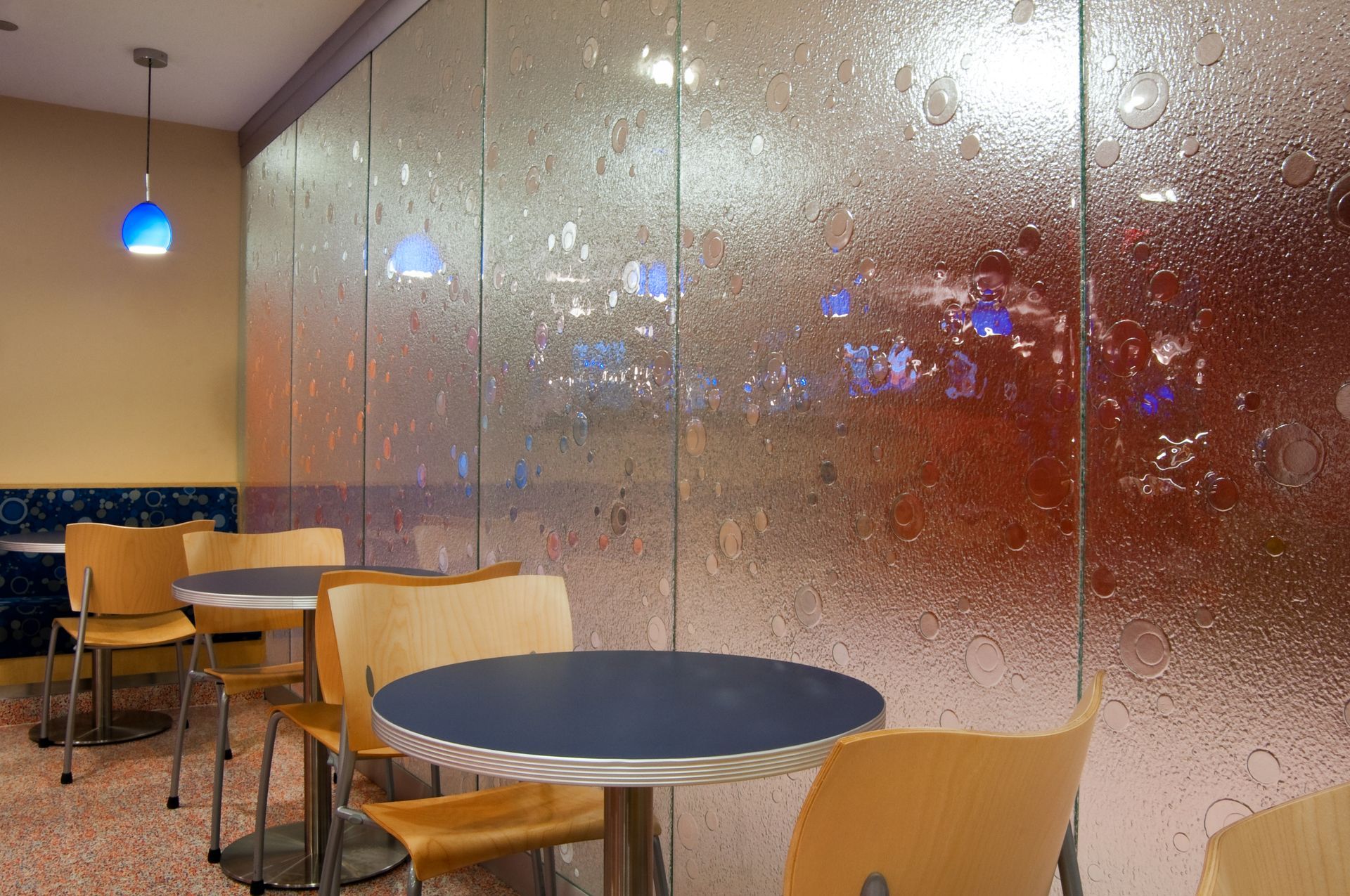 Empty café with round tables, wooden chairs, and a frosted glass wall with colorful lights beyond.