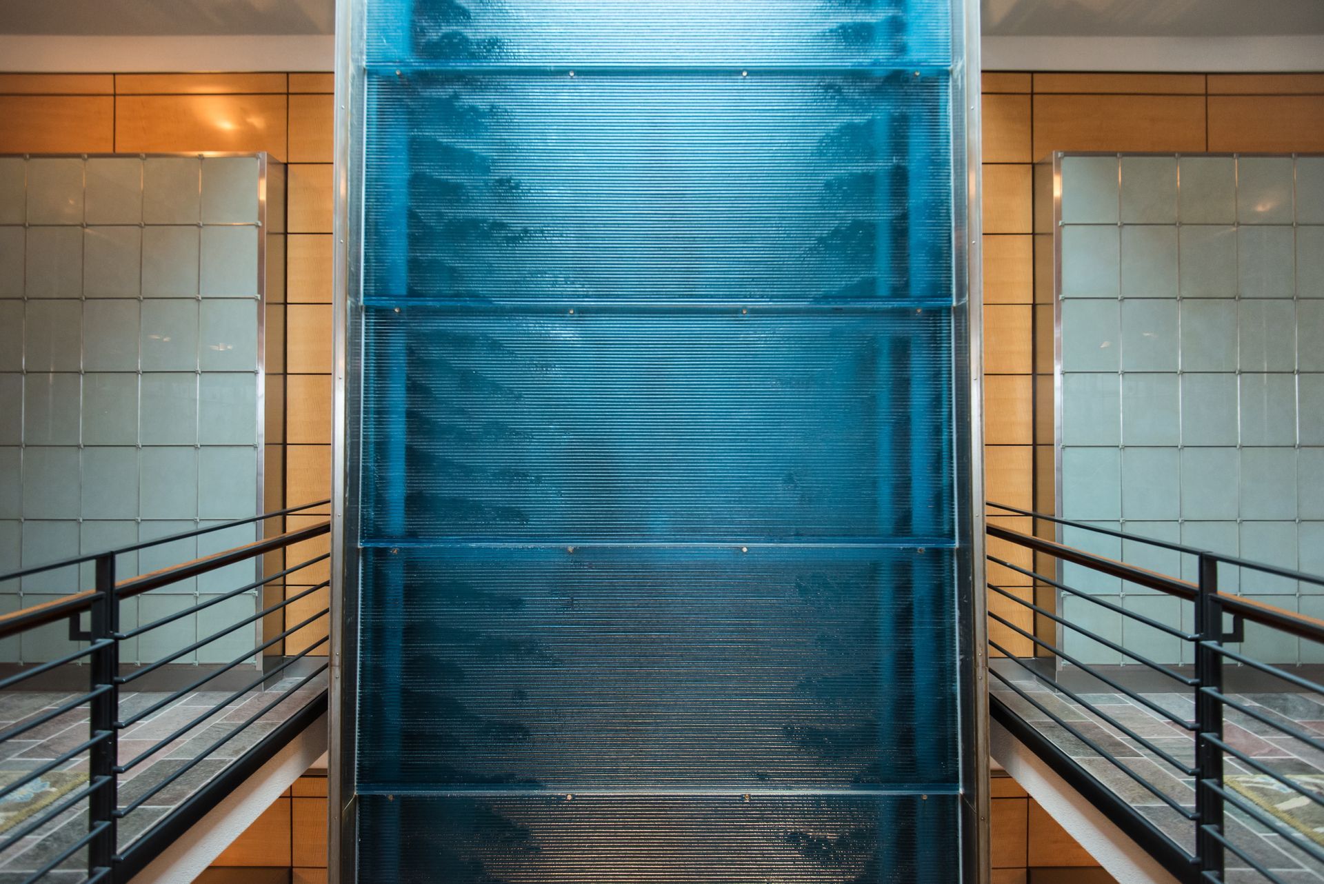 Blue glass elevator in a modern building hallway with metal railings and warm wall lights