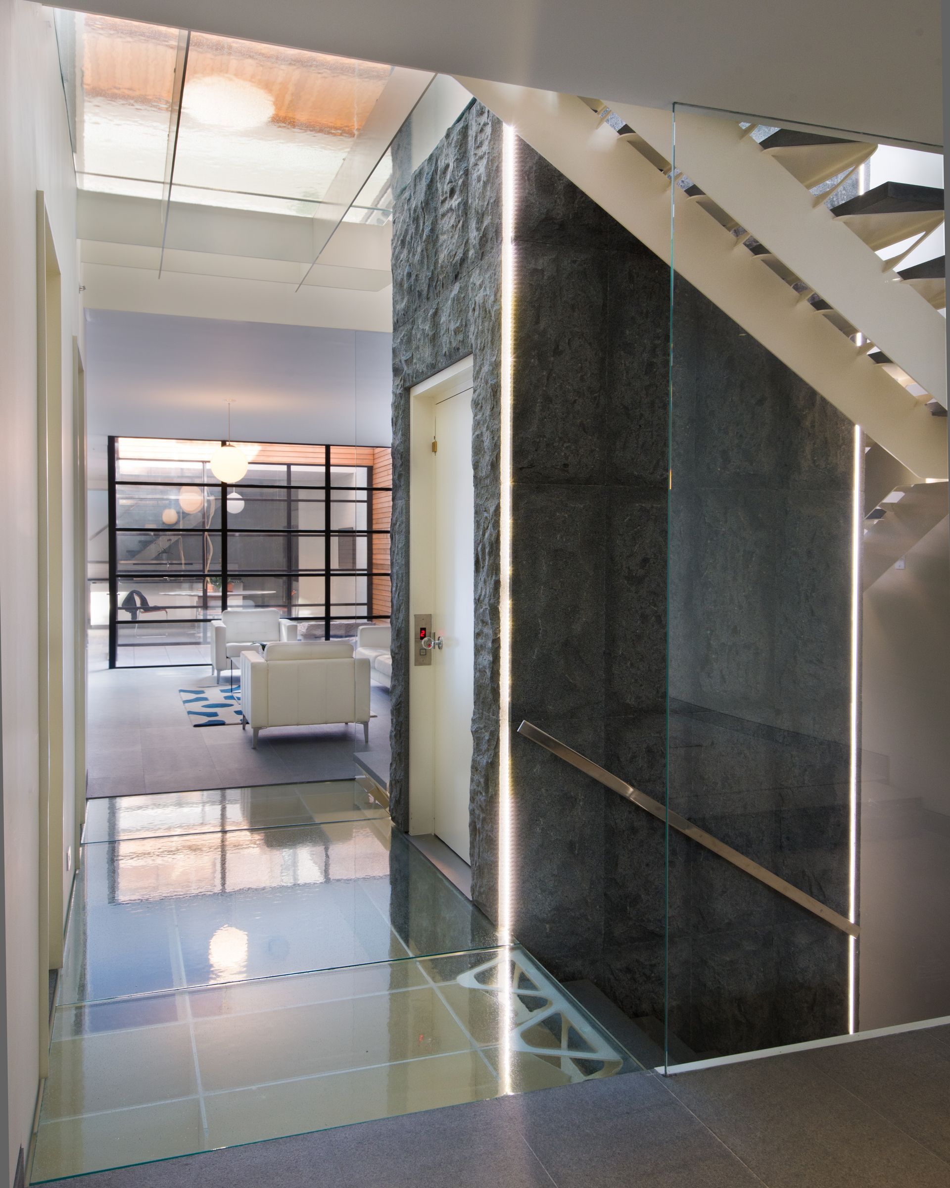 Modern office hallway with glossy tile floor, staircase, and a dark elevator shaft beside glass-walled rooms