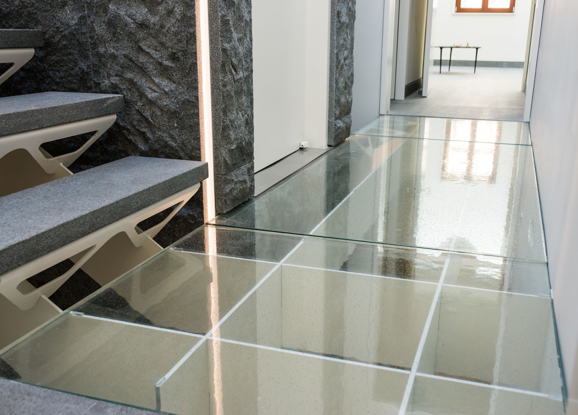 Glass-floored hallway beside a staircase, with stone walls and bright light reflecting on the floor