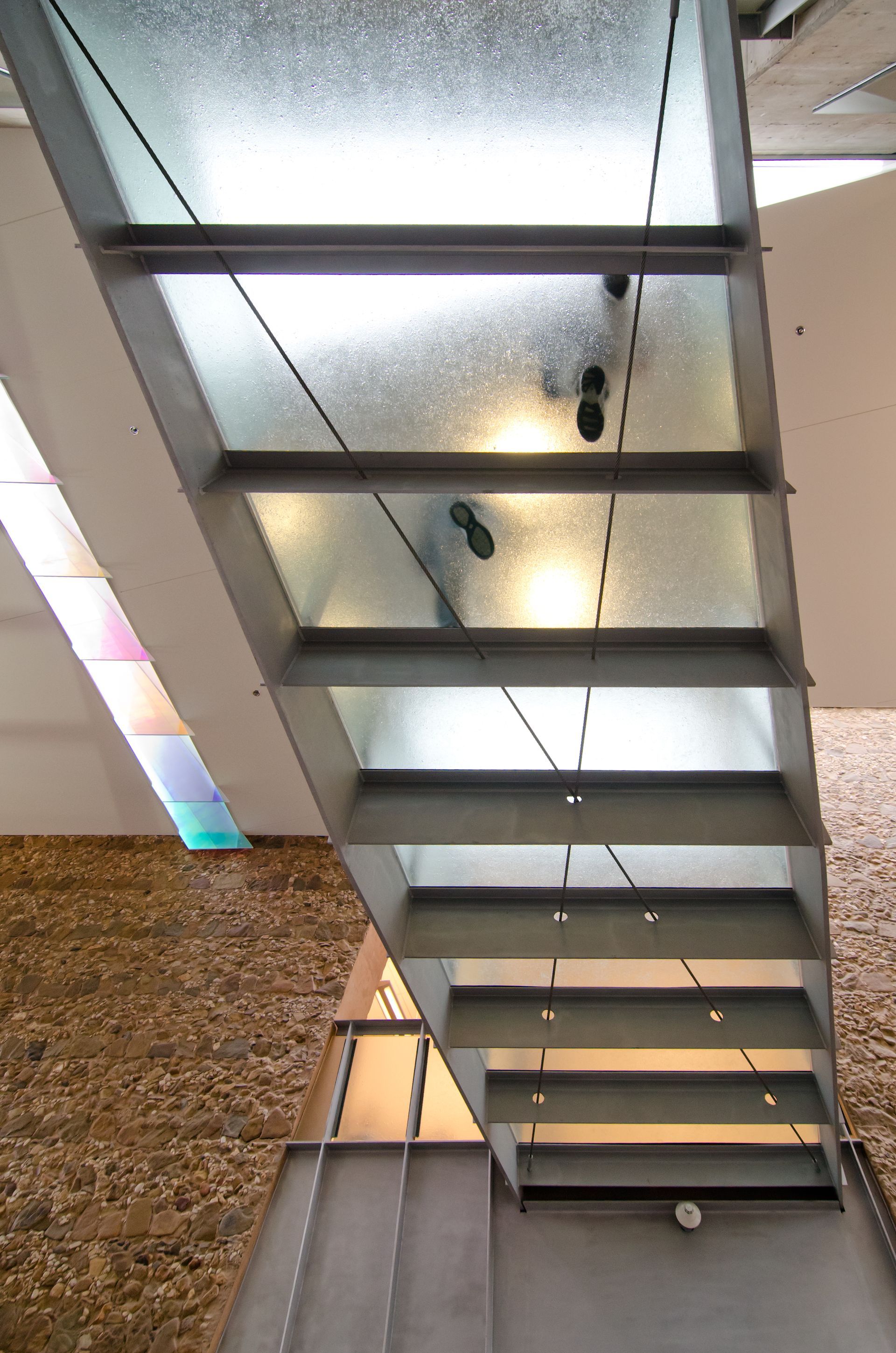 Underside view of a modern glass-and-metal staircase with lights and stone walls