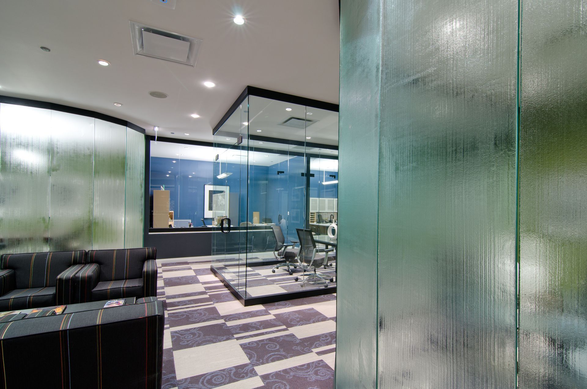 Modern office lobby with glass walls, patterned carpet, and black chairs under bright ceiling lights