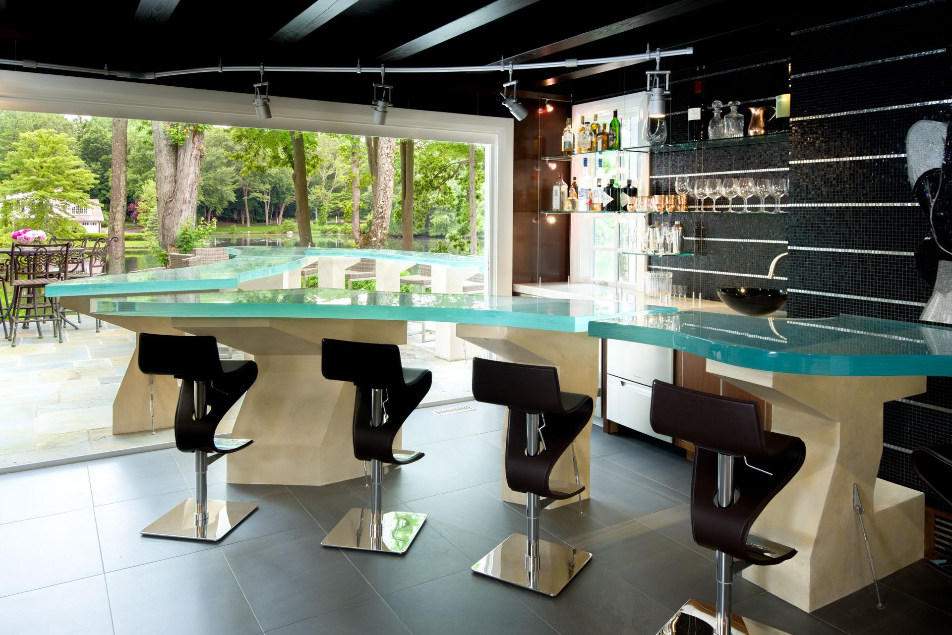 Modern bar interior with a turquoise countertop, black stools, and a view of greenery outside