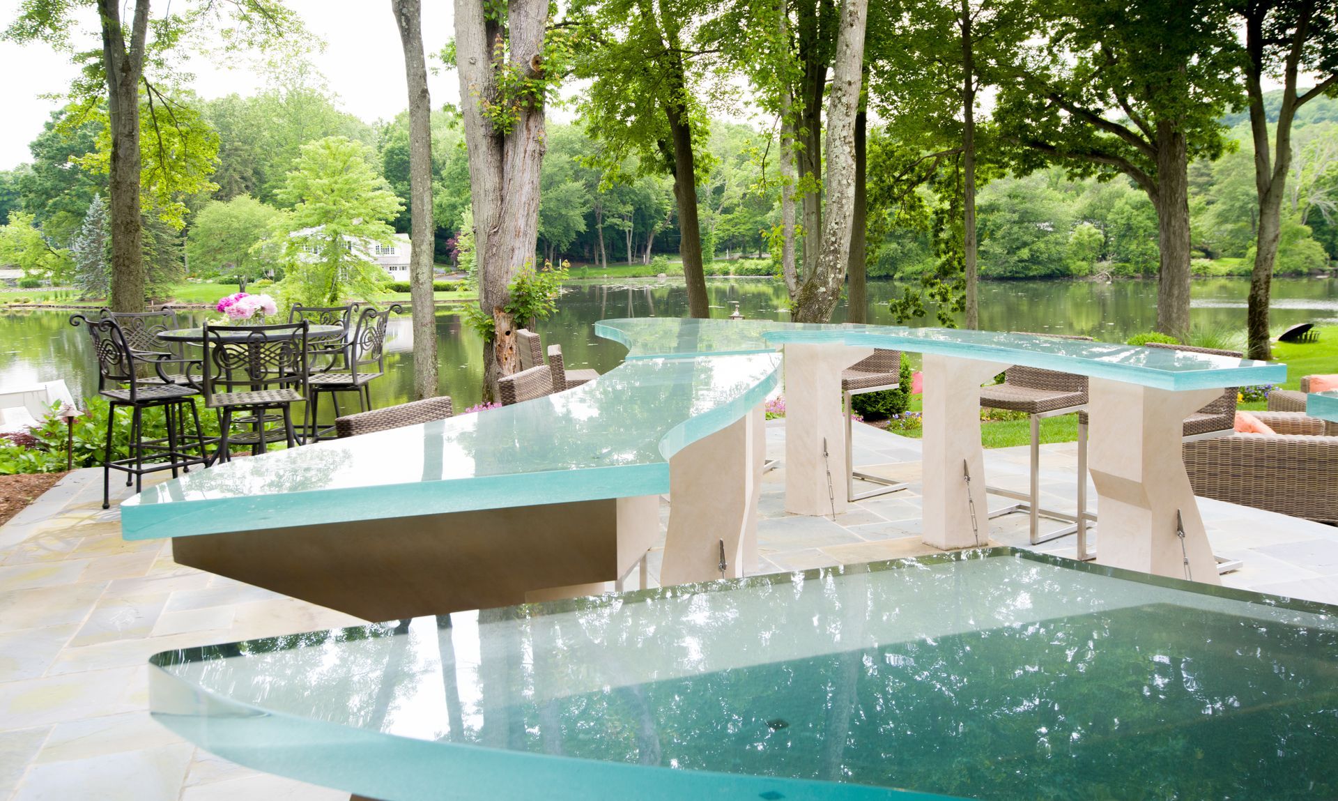 Modern outdoor poolside seating with curved glass tables and white chairs beside a garden pool