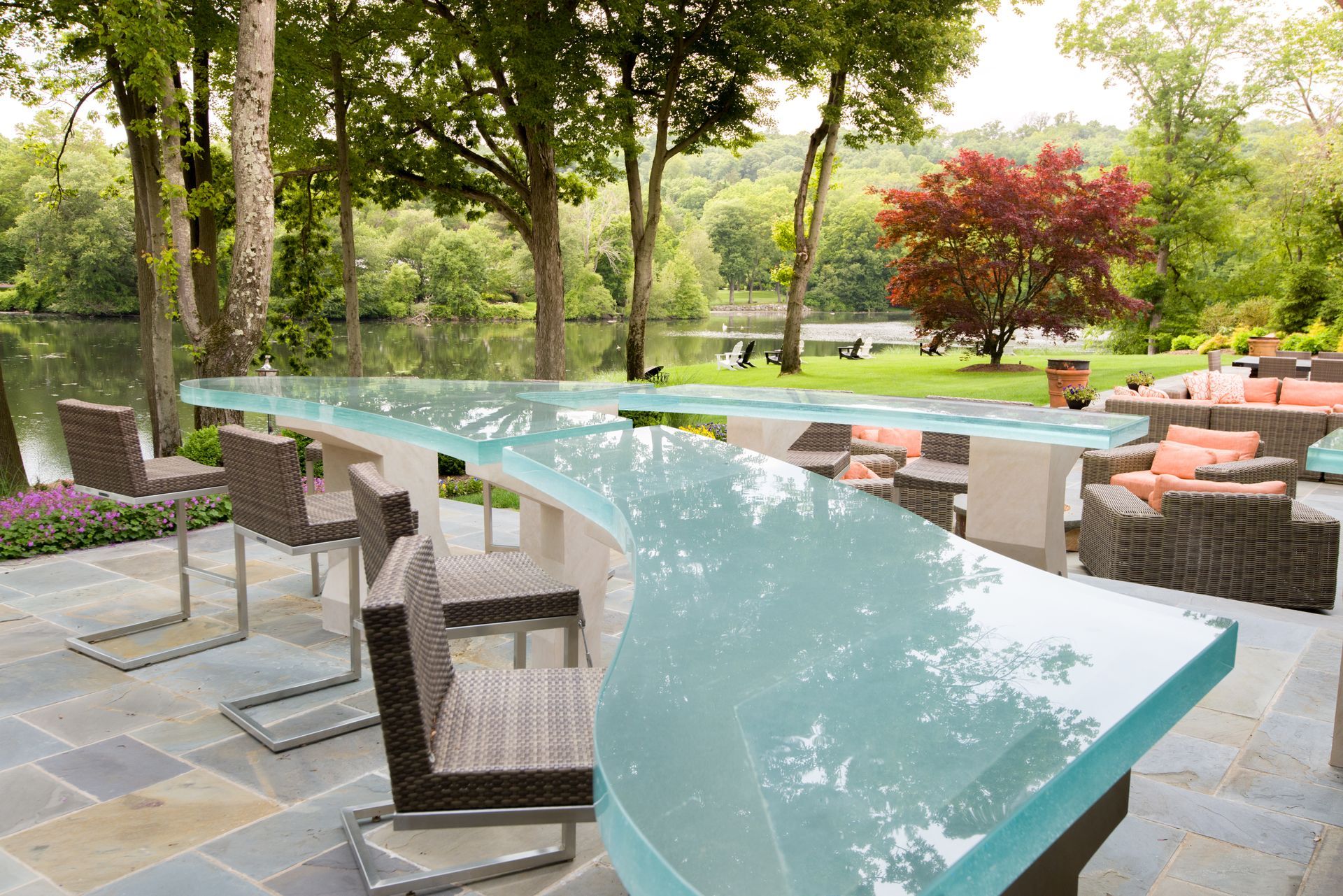 Curved glass patio table with wicker chairs on a garden terrace surrounded by trees and flowers
