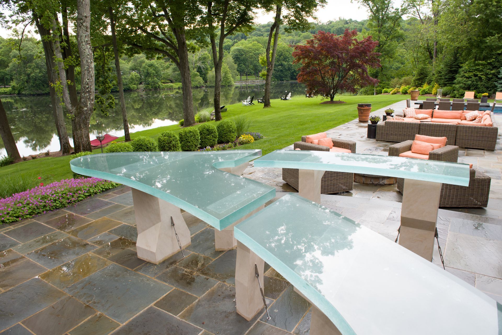 Modern glass outdoor table on a patio beside landscaped garden seating and trees