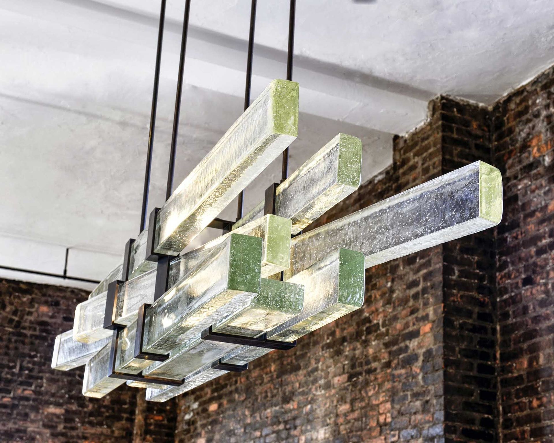 A chandelier made of cast glass mouldings  hanging from the ceiling in front of a brick wall.